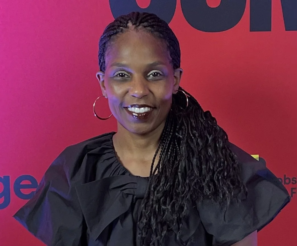 A woman with braided hair, hoop earrings, and dark lipstick smiling at the camera, standing in front of a red background with black text.