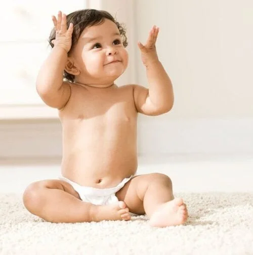 A baby sitting on a carpet with arms raised and a curious expression.