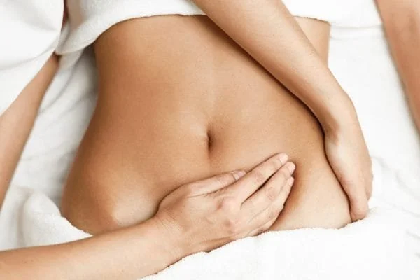 A close-up of a person's bare abdomen with hands gently pressing on the stomach, lying on a white surface.