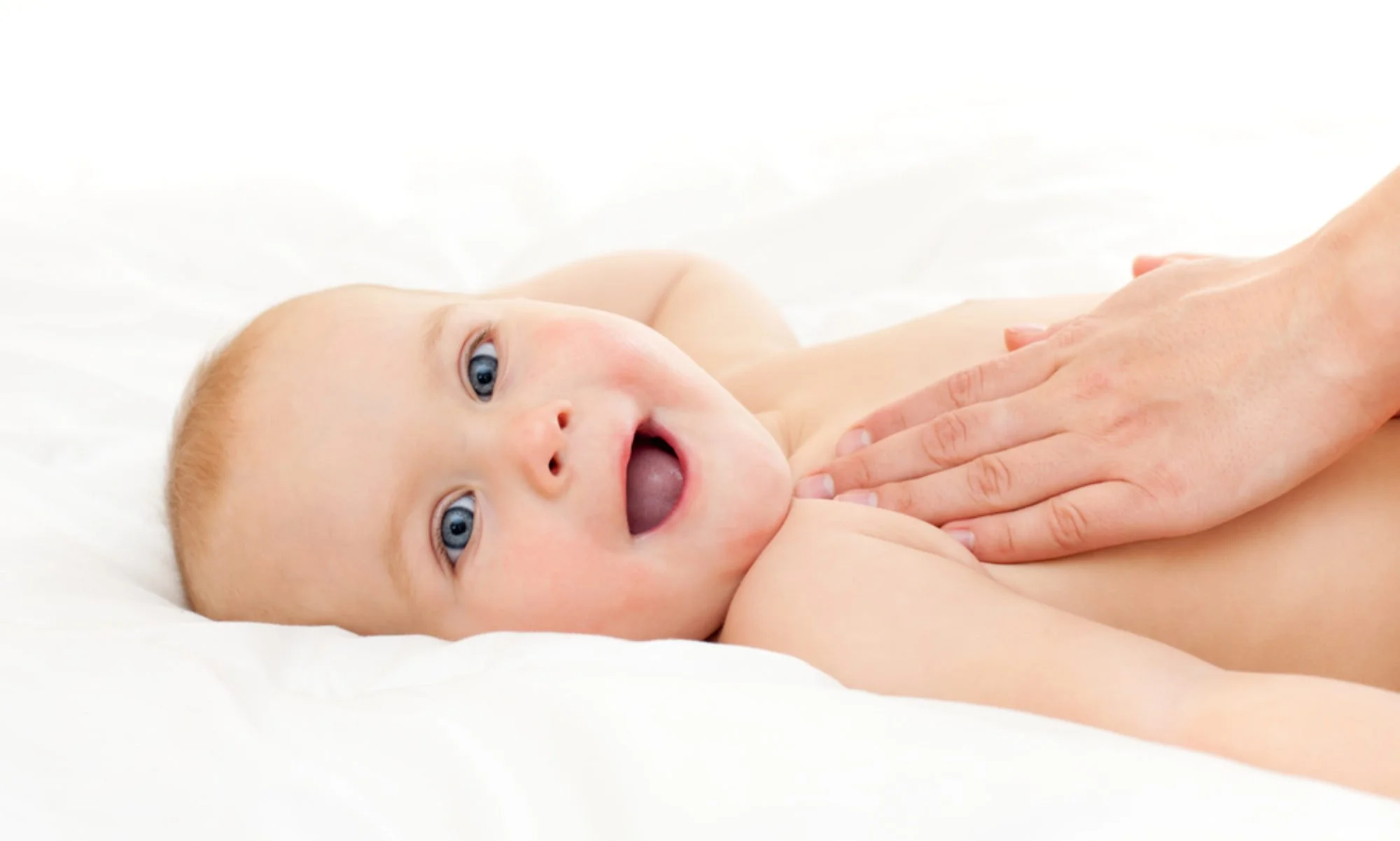 A baby lying on a white surface, with an adult's hand gently touching its chest, looking at the camera with wide blue eyes and an open mouth.