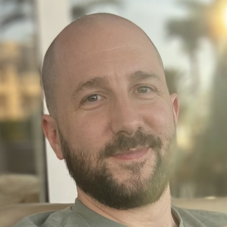 Ein lächelnder Mann mit Bart und kurzhaarigem Kopf, im Hintergrund eine Fensteransicht mit natürlichem Licht.