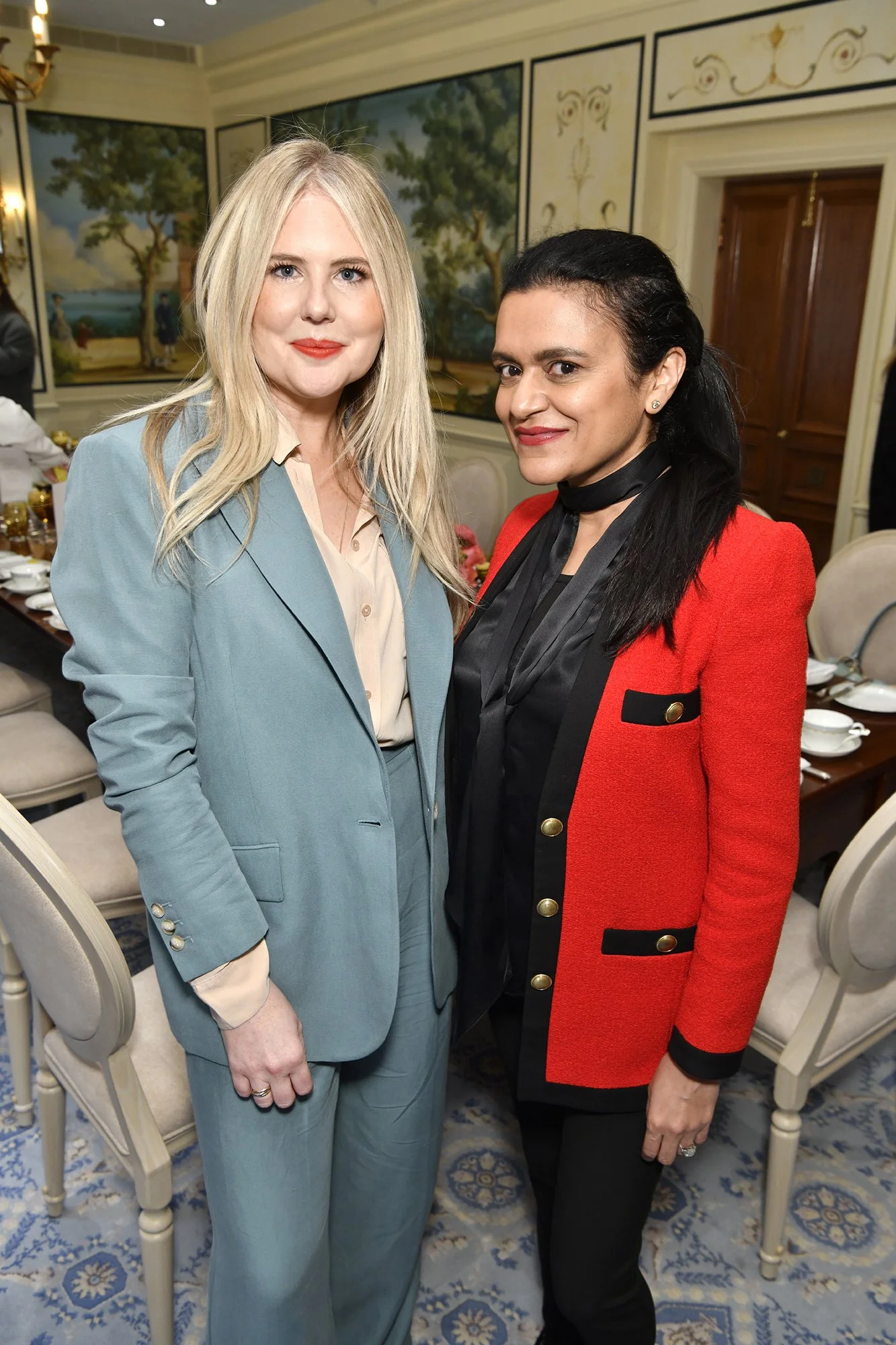 Two women standing in a dining room, smiling at the camera. The woman on the left has long blonde hair and is wearing a light blue suit. The woman on the right has dark hair pulled back and is wearing a red blazer with gold buttons and black trim, and a black scarf. The background features paintings of trees and a decorated table set for a meal.