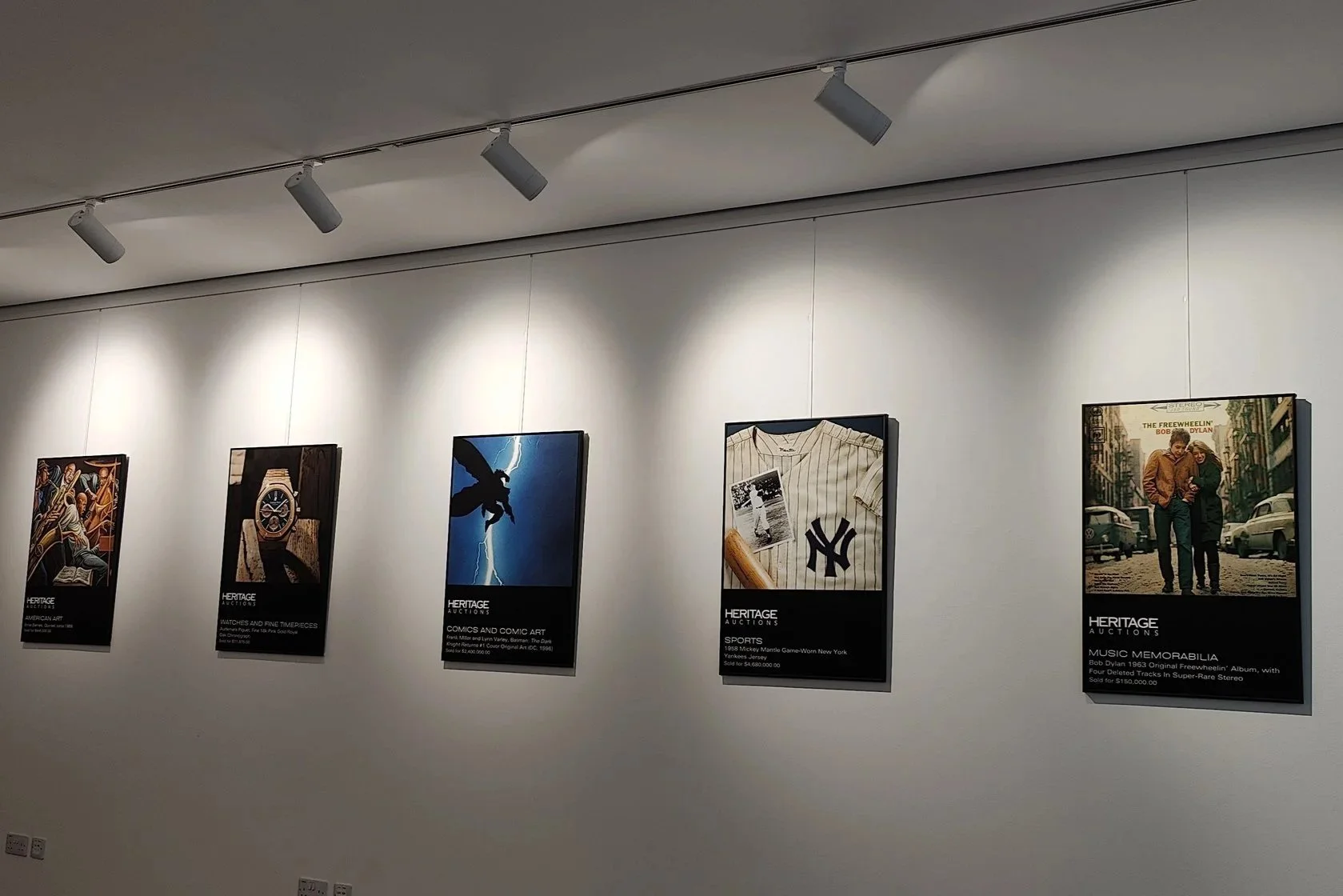 Gallery wall with five framed posters, each with black labels underneath, in an art exhibition space, illuminated by ceiling track lighting.