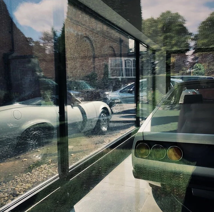 Reflection of several cars parked outside viewed through a large glass window, with part of an interior vehicle visible inside.