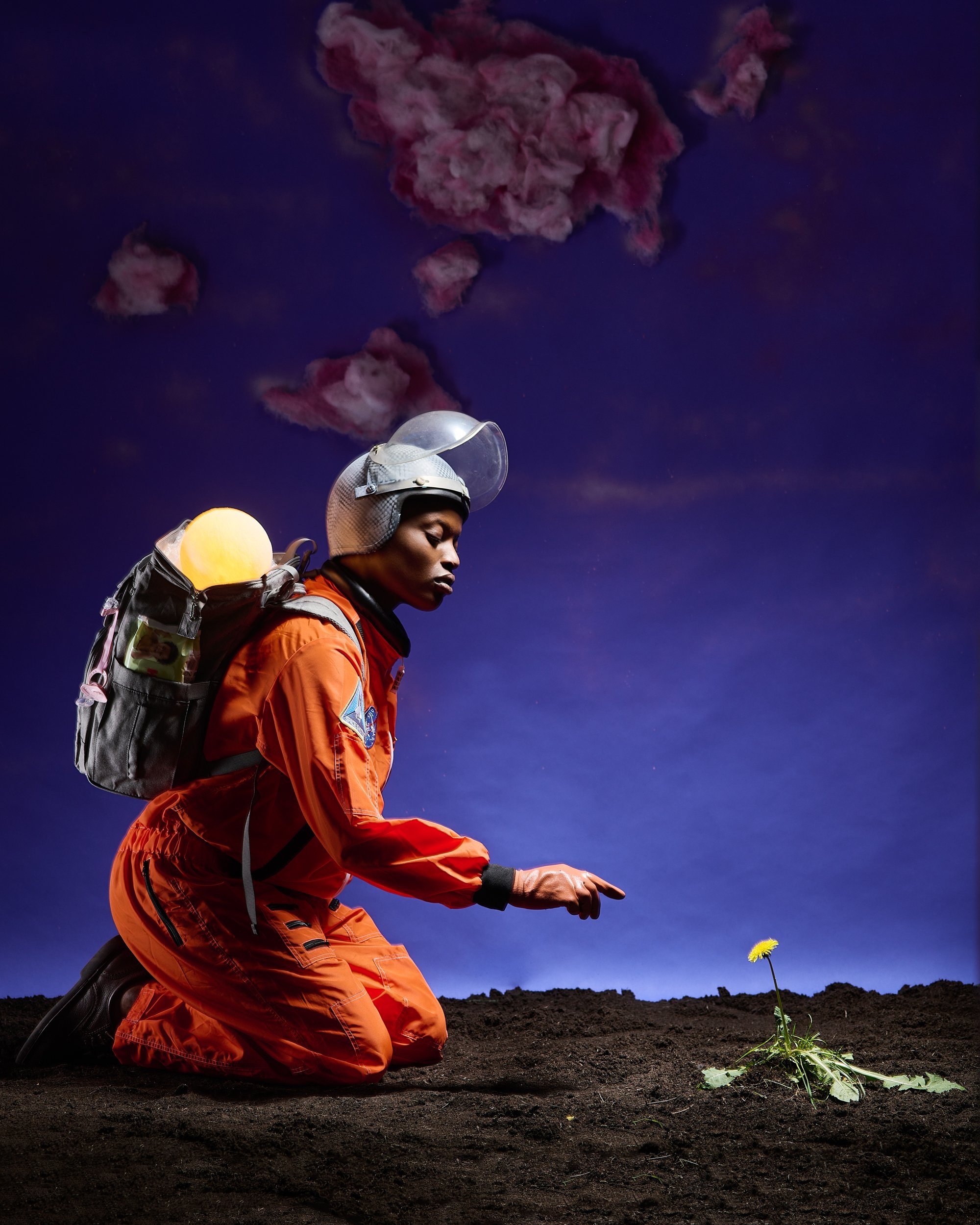 Person in an orange jumpsuit kneeling on dirt ground, pointing at a small yellow flower. The person is wearing a helmet with a clear visor, a backpack with a glowing yellow light, and is set against a dark sky with pink clouds.