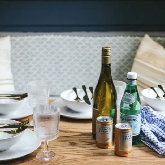 Table set for a meal with white bowls, metal utensils, glasses, a wine bottle, a water bottle, and canned drinks on a wooden table.