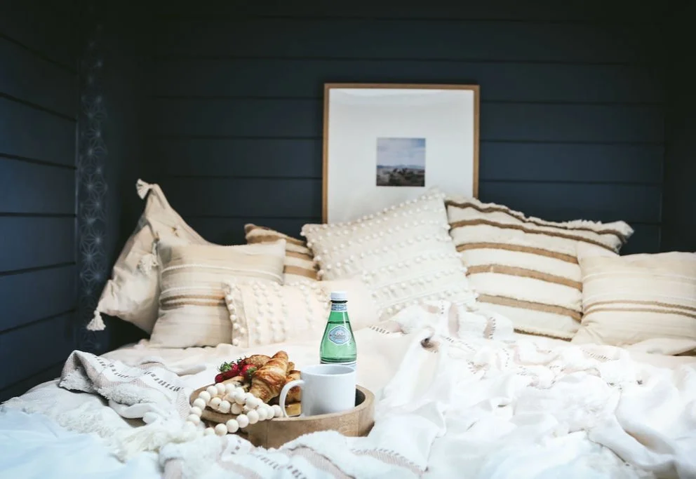 A cozy bed with multiple cream-colored pillows, a tray with a bottle of sparkling water, a mug, and a croissant, in front of a framed picture hanging on a dark blue wooden-paneled wall.