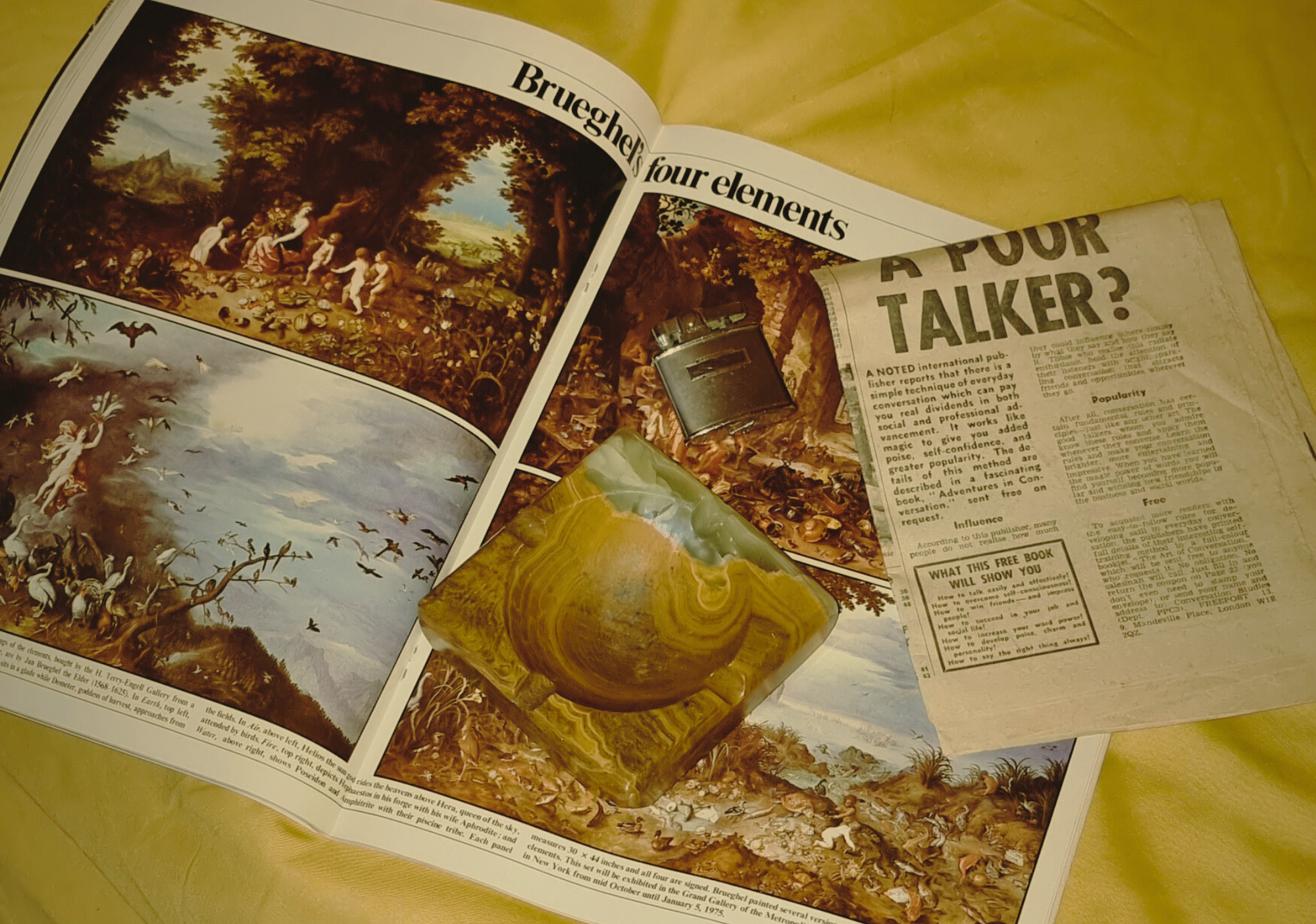 Open art book displaying classic paintings of nature scenes, a newspaper titled 'A POOR TALKER?' with printed text, a small black object, and a polished stone with swirling colors on a yellow surface.