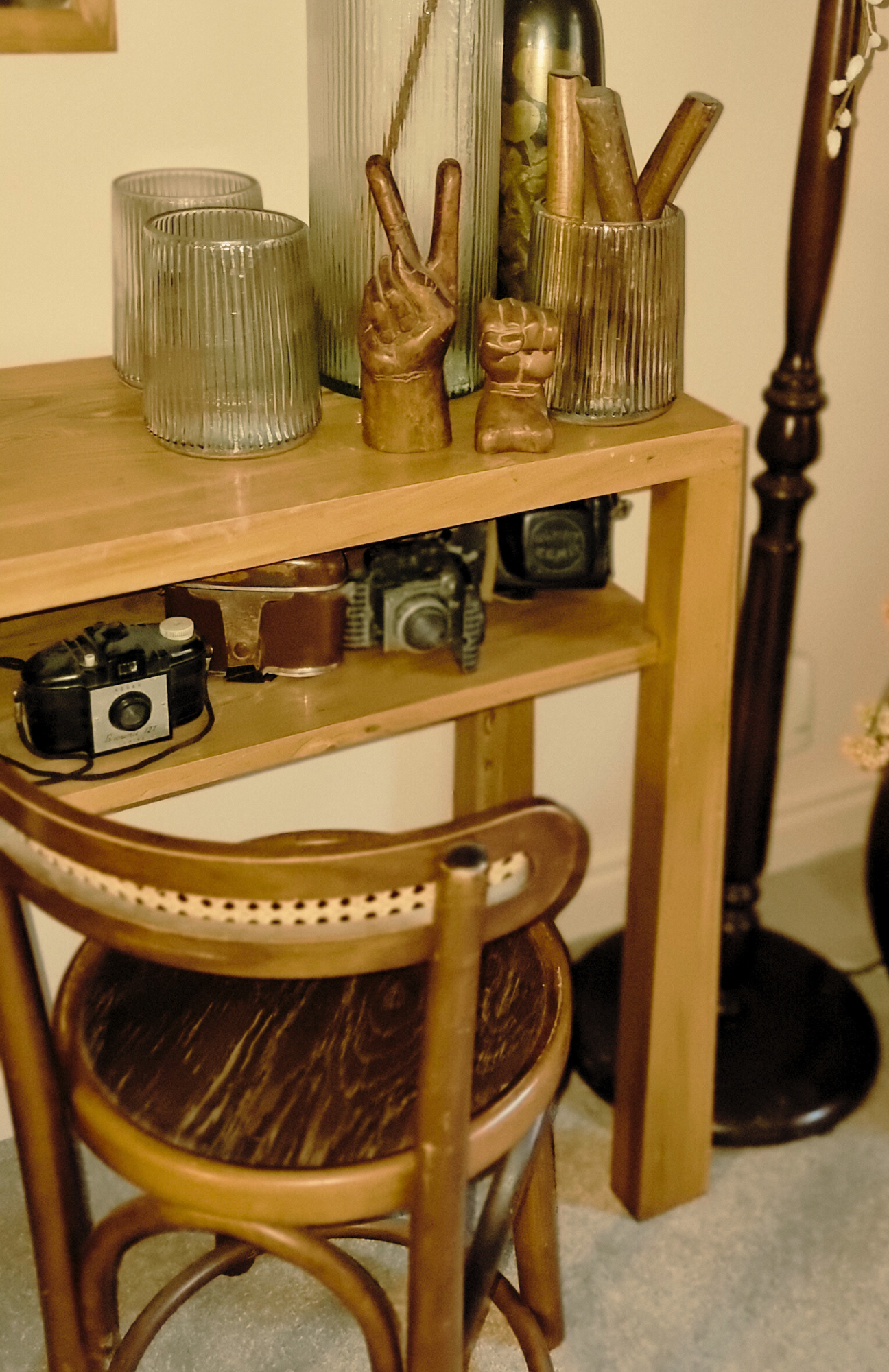 A wooden table with decorative items including glass cups, wooden hands making peace and fist gestures, and books, with two vintage cameras and a wooden chair in the foreground.
