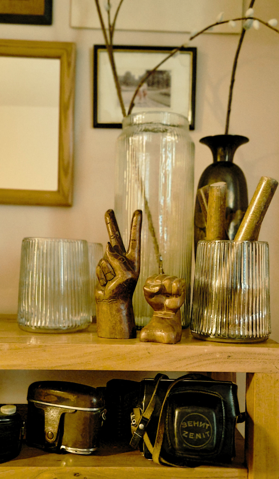 Decorative shelf with two glass vases, one tall and ribbed, the other shorter and ribbed, a gold-colored sculpture of a peace sign, and a fist. Below, on the shelf, there are various camera and camera-related items. In the background, framed photogra