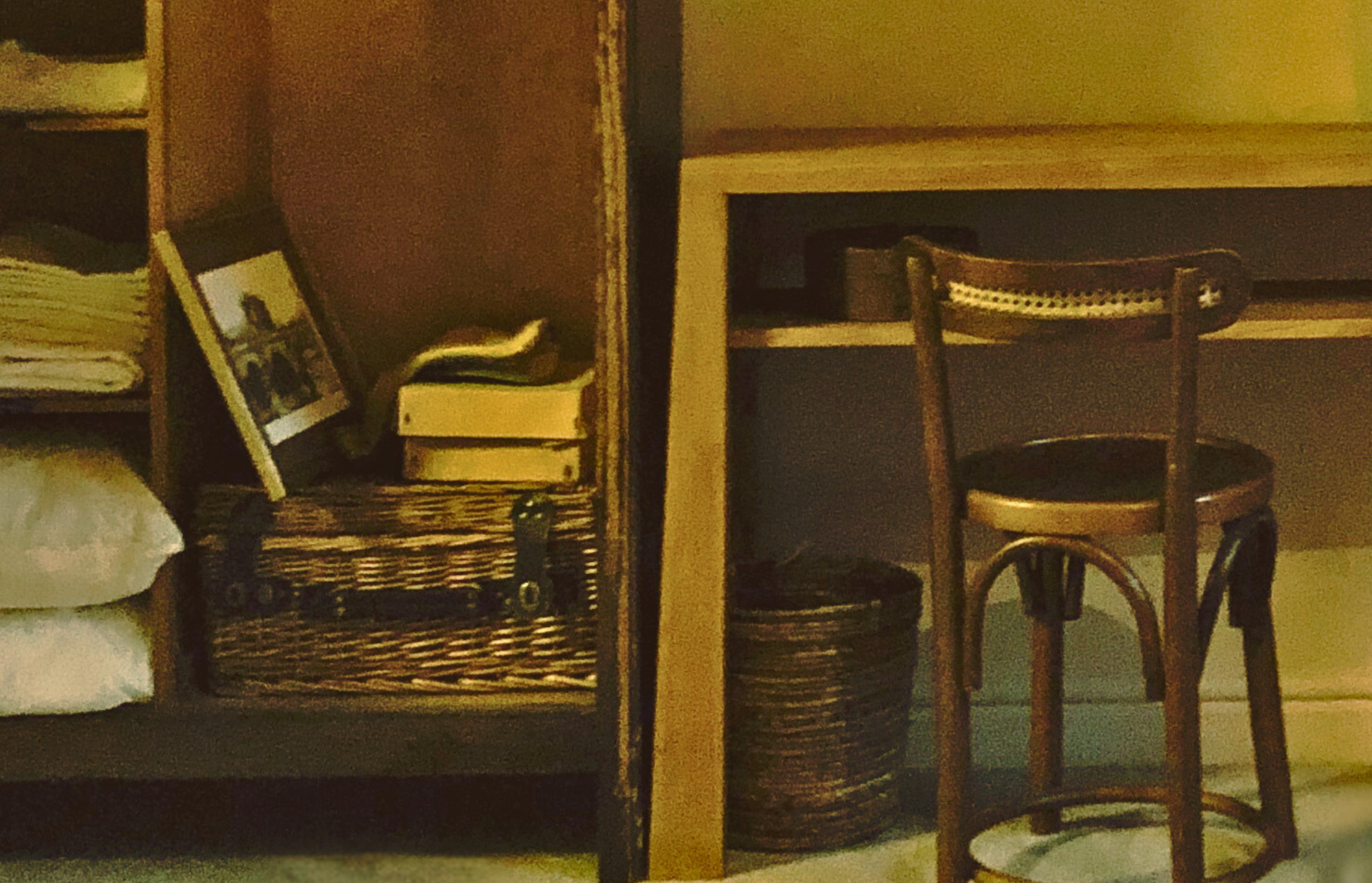 Interior scene with wooden shelves, framed photograph, books, and a woven basket on the left; small table, wooden chair, and storage basket on the right.