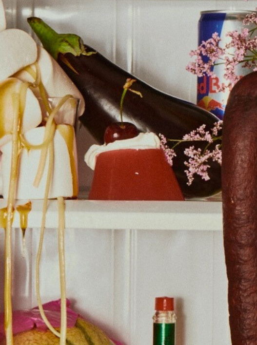 A shelf with an eggplant, a cherry on top of a dessert, a can of Red Bull energy drink, pink flowers, and a partially visible bottle.