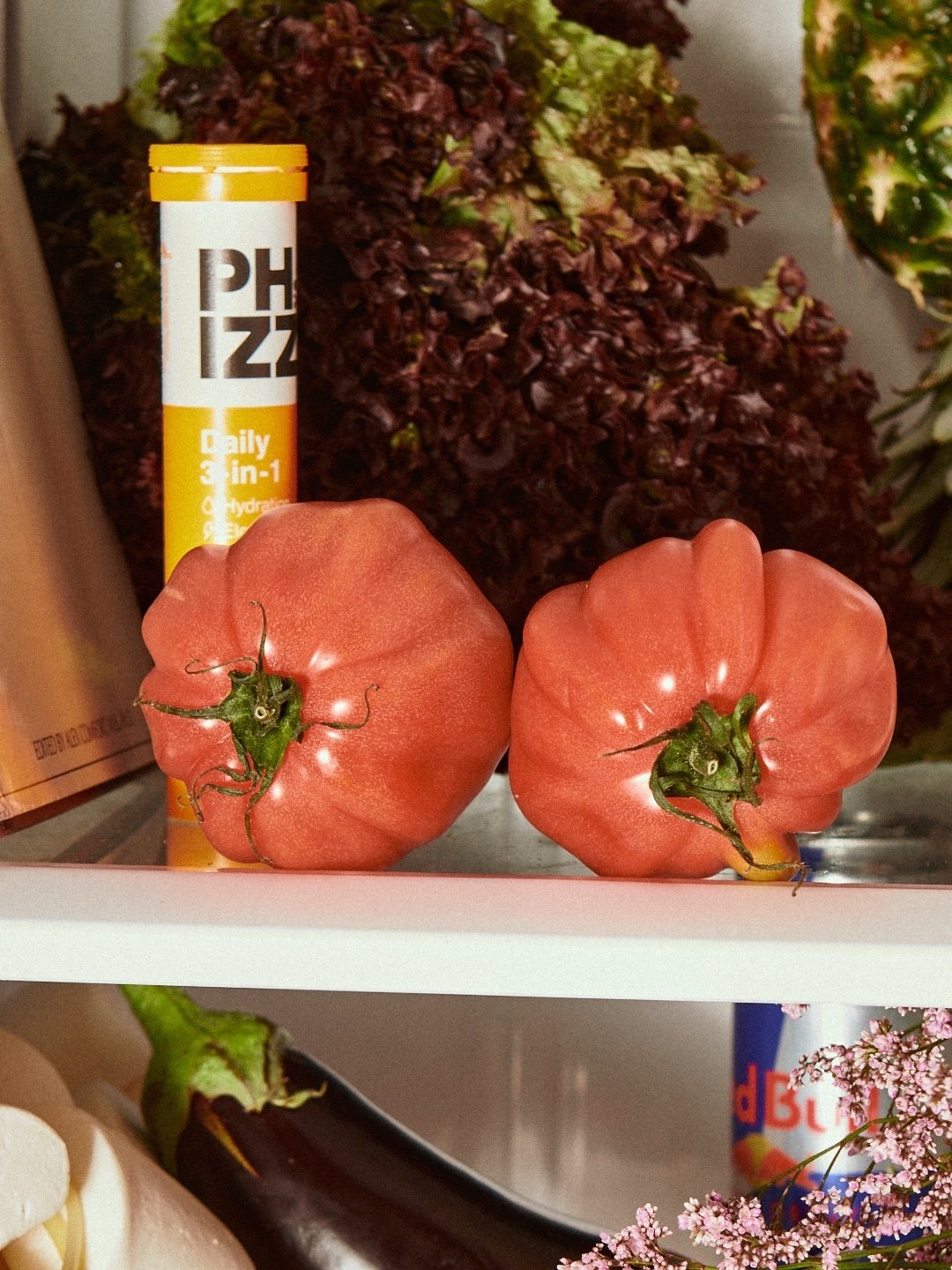 Two heirloom tomatoes with irregular shapes and green stems, placed on a shelf next to a tube and purple leafy lettuce in the background.