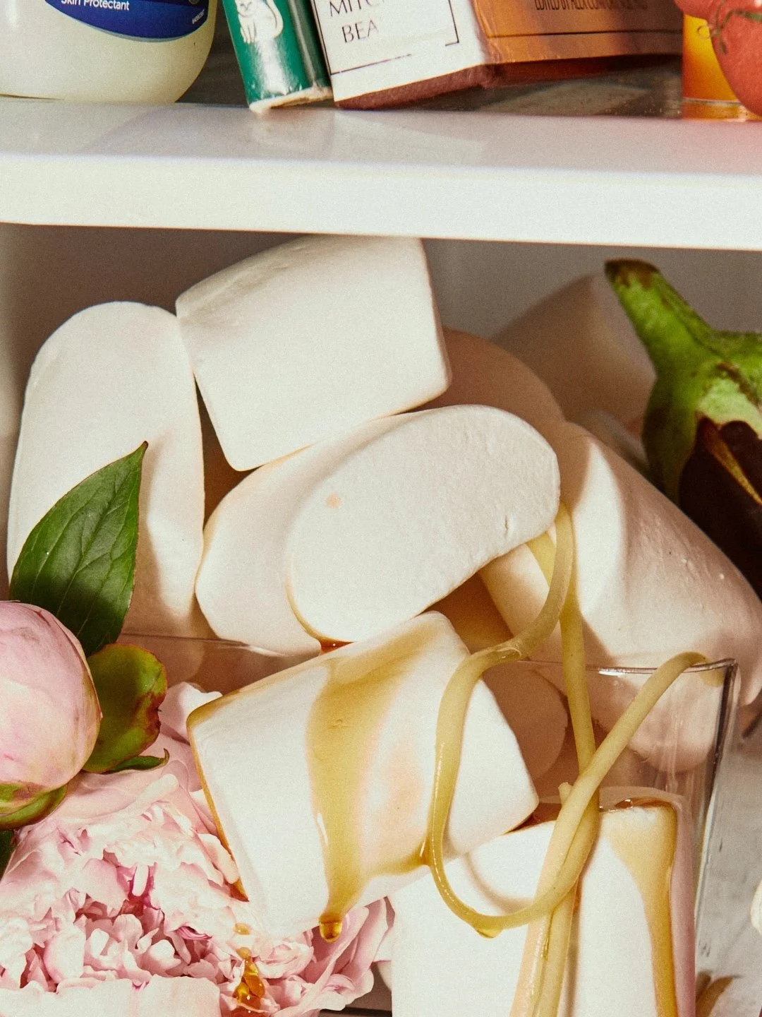 Marshmallows with honey drizzled over them, pink flowers, a green leaf, and a part of an eggplant in a clear container.