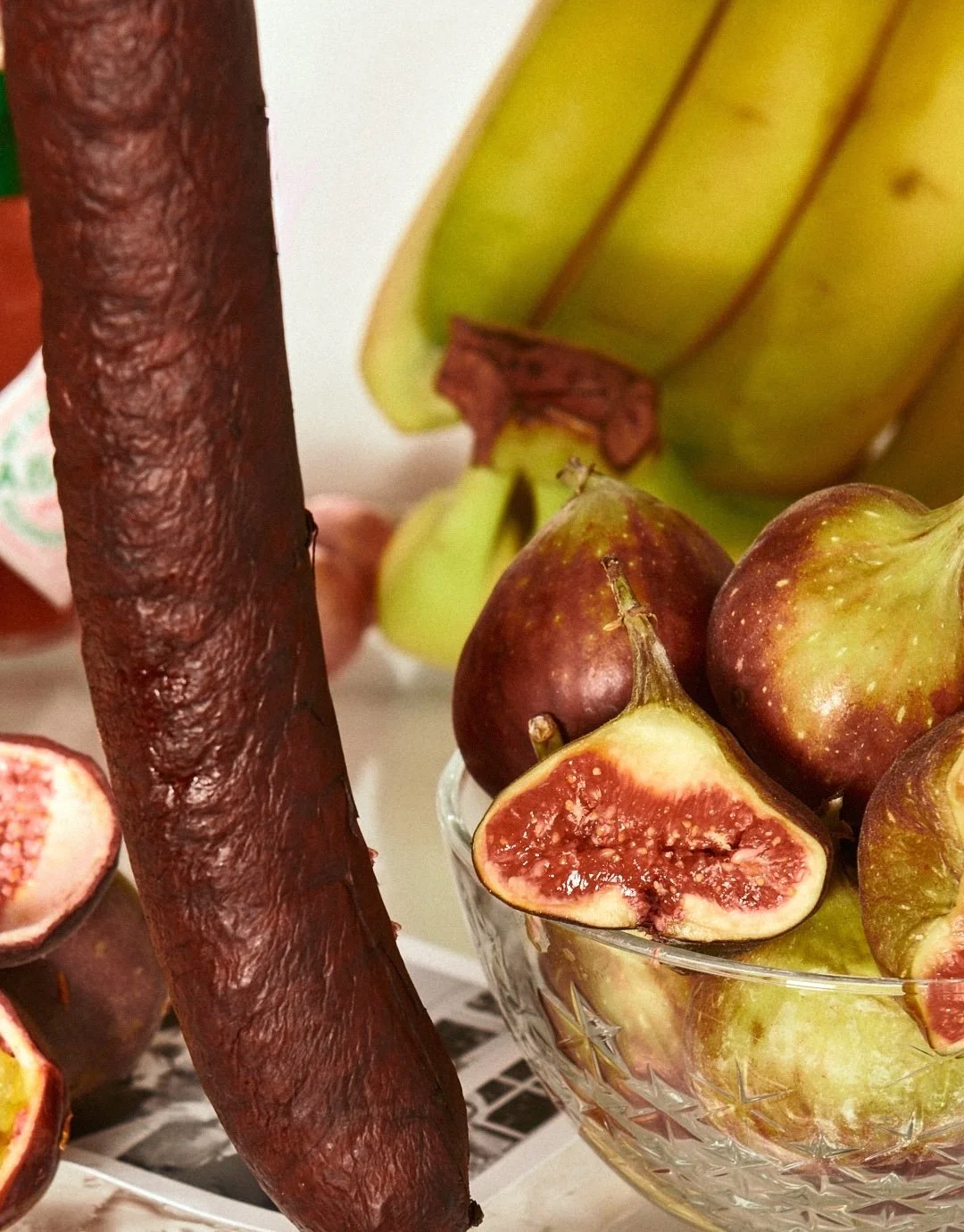 Close-up photo of a bowl of fresh figs with some halved showing their red interior, bananas in the background, and a rimmed chocolate or cacao bar on the left side.