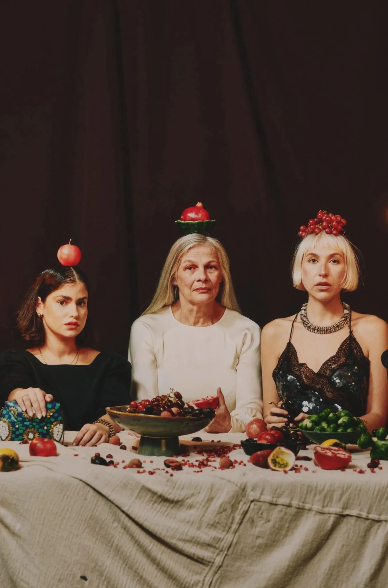 Three women sitting at a table with fruit, each with fruit on their head. The woman in the middle has a pomegranate on her head, the woman on the left has an apple, and the woman on the right has a bunch of grapes.