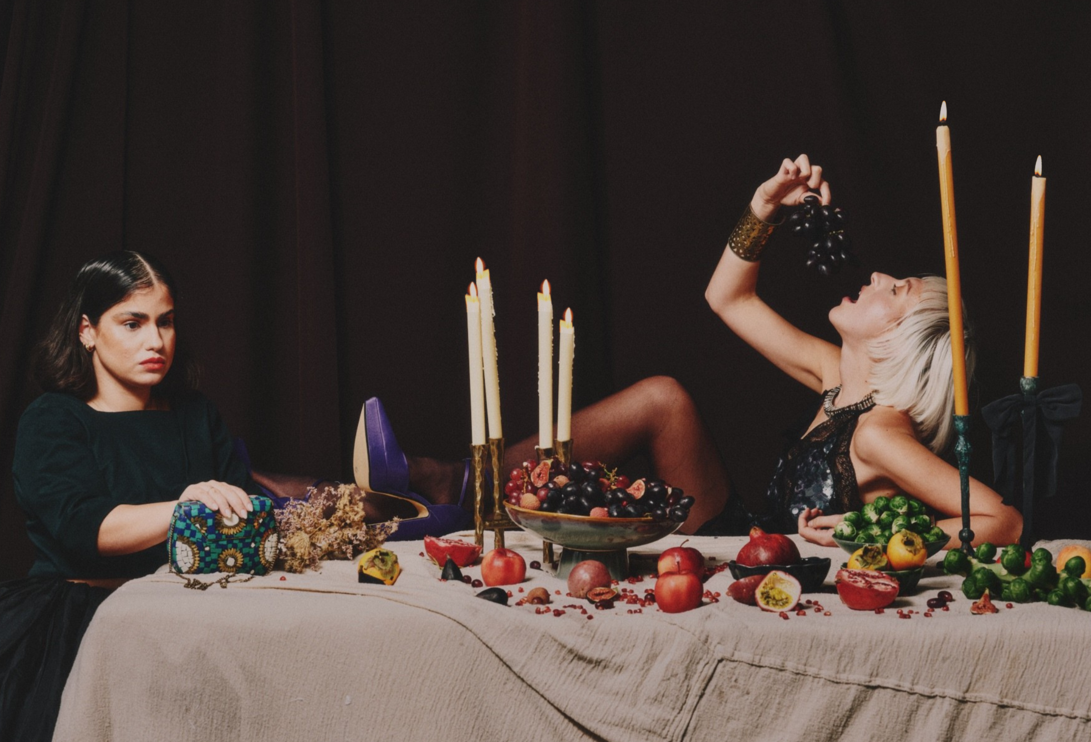 Two women sit at a table with fruit and candles. The woman on the left looks serious, while the woman on the right reclines, holding a bunch of grapes.