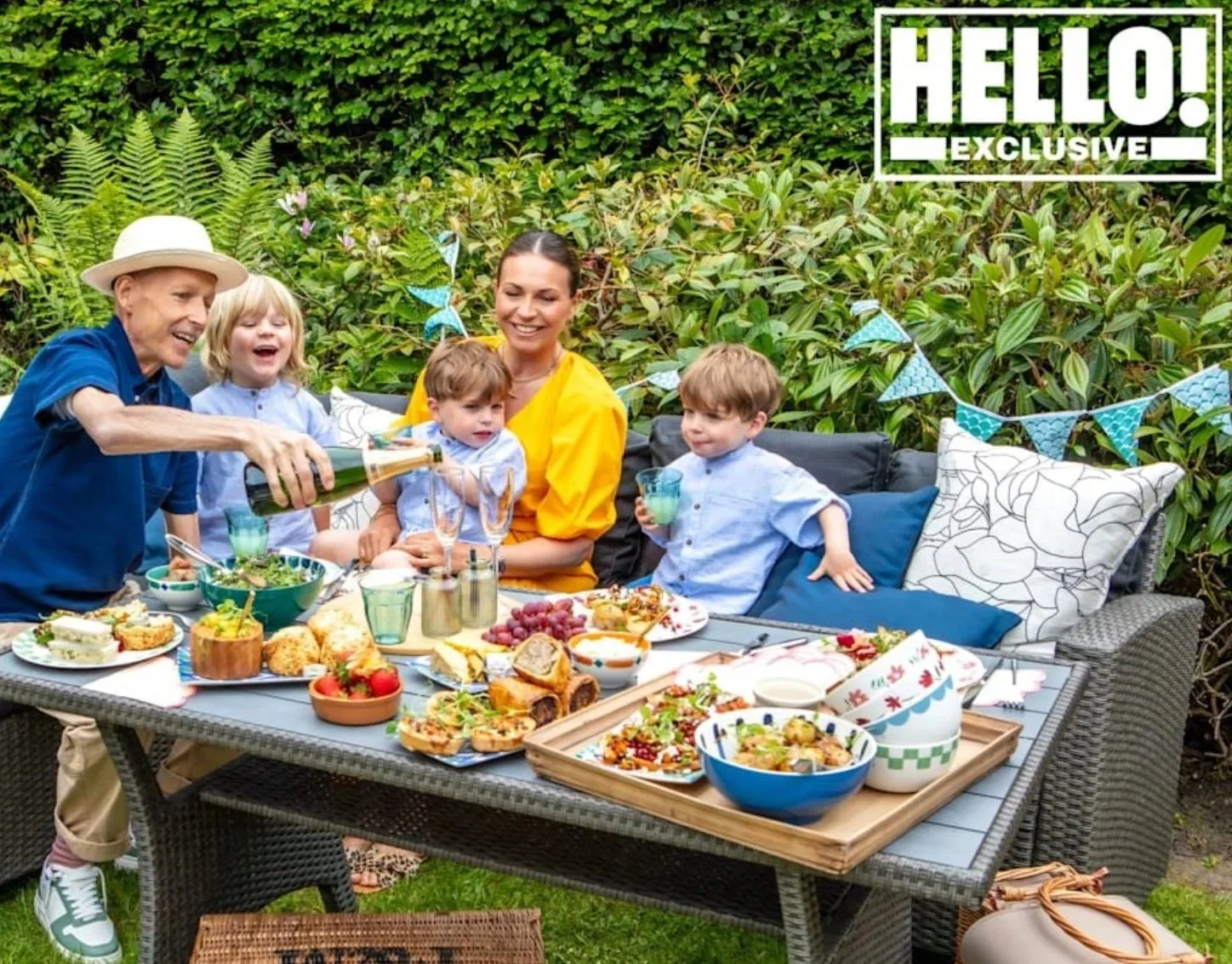 Family gathered around outdoor dining table with food, drinks, and decorations, celebrating in a garden.
