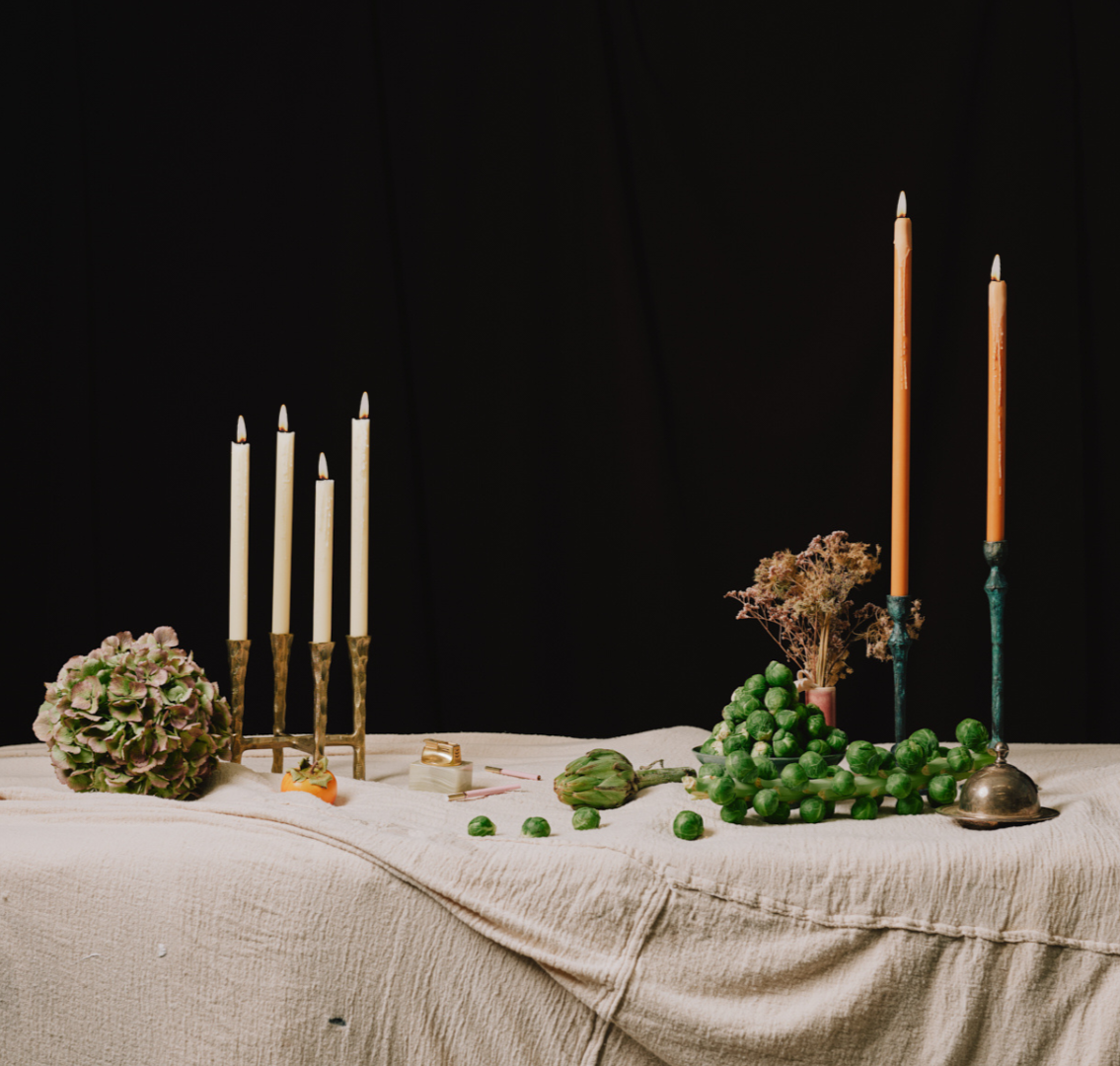 An elegant table setting with a beige tablecloth, decorated with lit candles, green Brussels sprouts, artichokes, dried flowers, and decorative objects, against a black background.