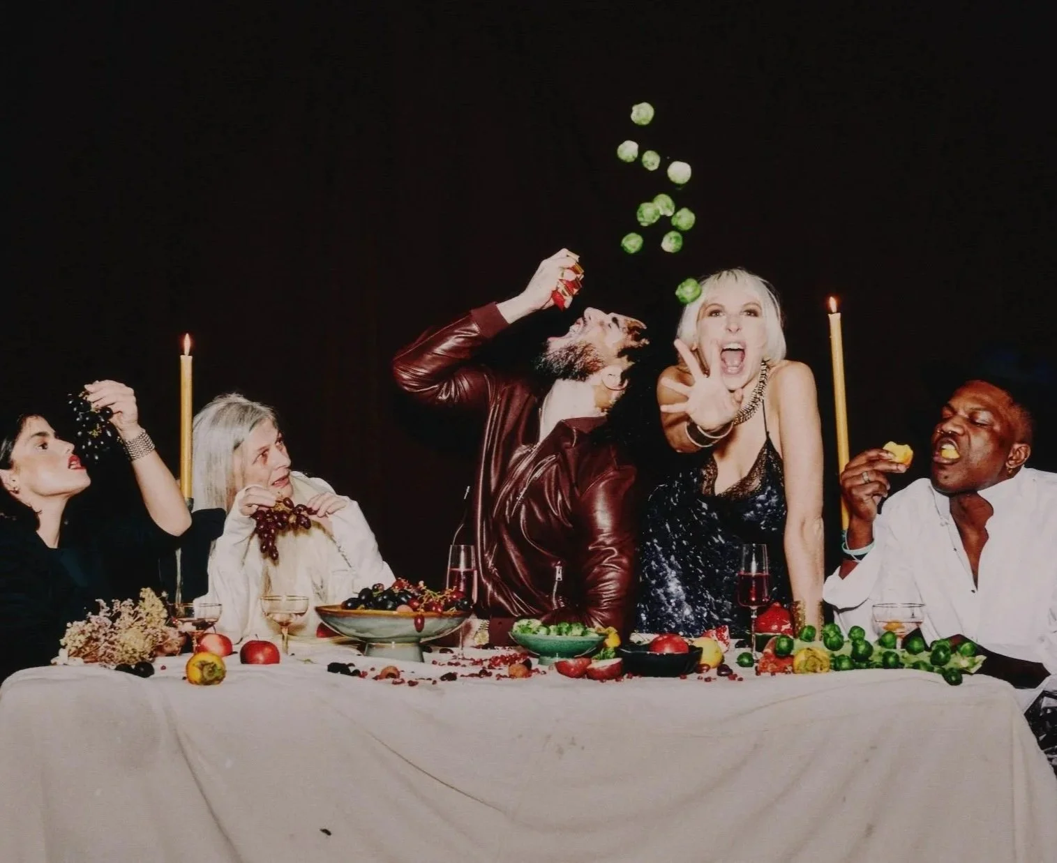 People celebrating at a dinner party with food, drinks, and candles, throwing grapes into the air in a lively and joyful scene.