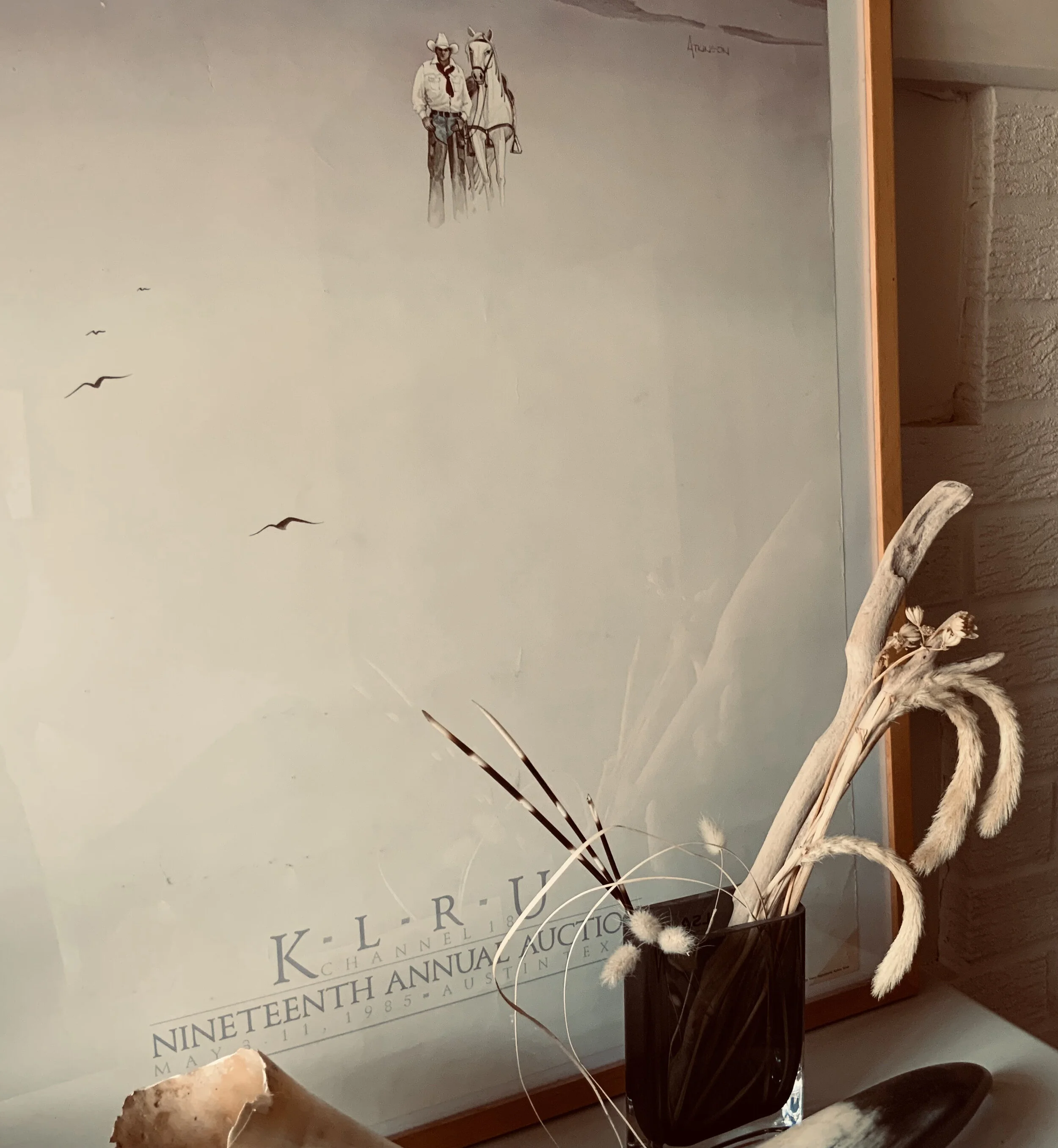 A framed poster or print leaning against a wall, featuring an illustrated cowboy with a hat, red bandana, and a white horse, with birds flying in the sky and some dried plants or decorative items in a black vase in the foreground.
