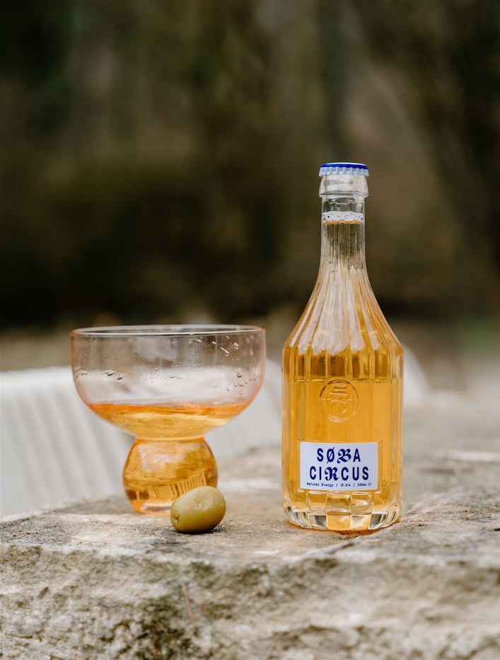 A bottle labeled 'Soba Circus' filled with a golden liquid, next to a matching bowl with liquid and an olive on a stone surface outdoors.