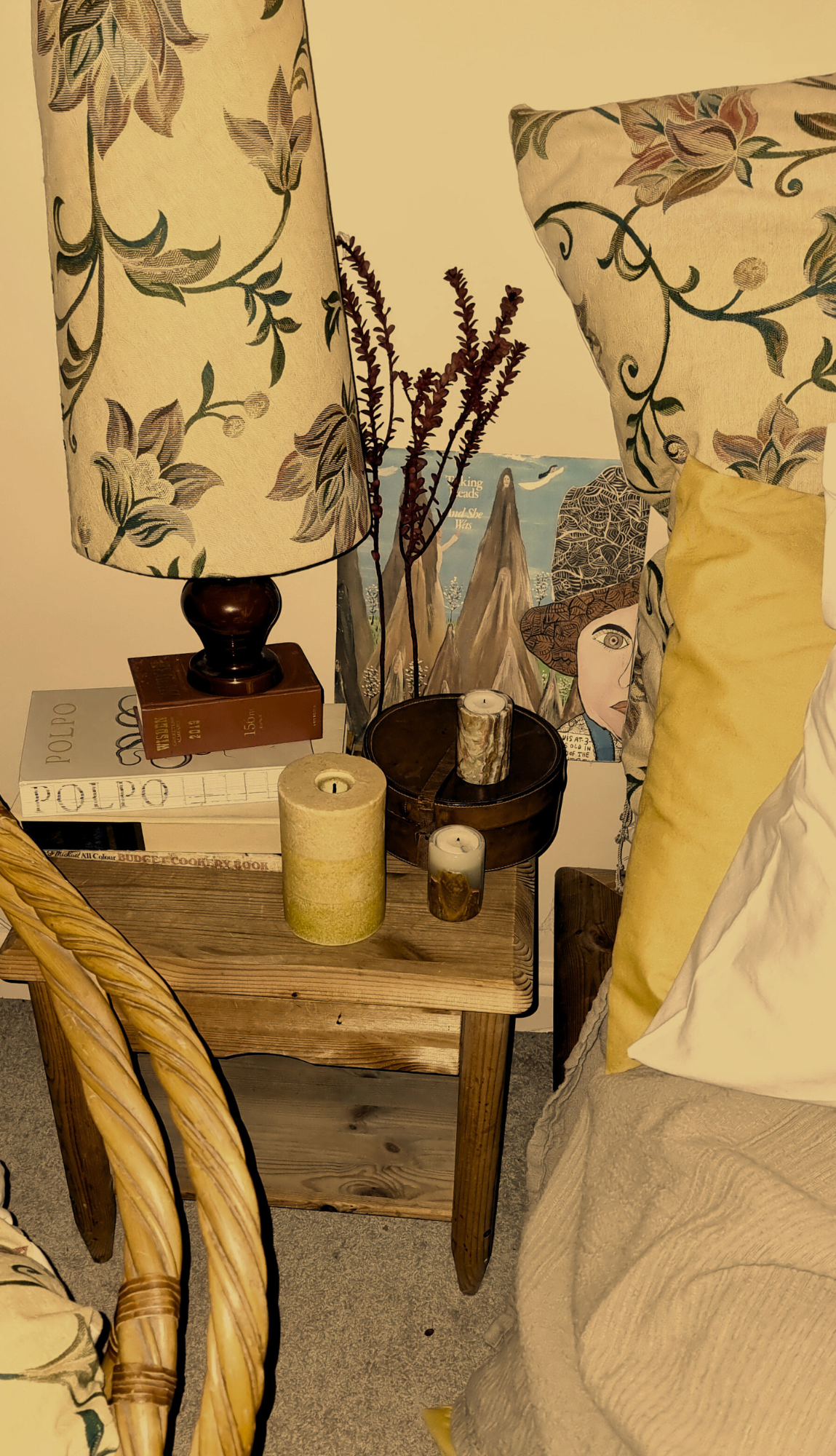 A cozy corner with a wooden side table holding candles, books, and decorative items, next to a bed with floral and yellow pillows, and a bed lamp with a floral lampshade.
