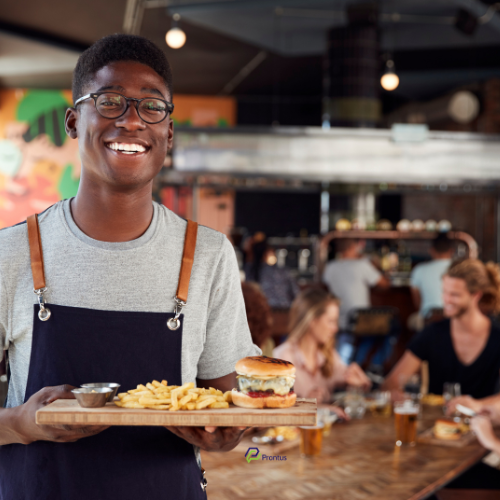 a male waiter in a grey tshirt and dark blue apron smiling as he carries a board with two sauce post, fries and a burger in a bun with cheese oozing out of it