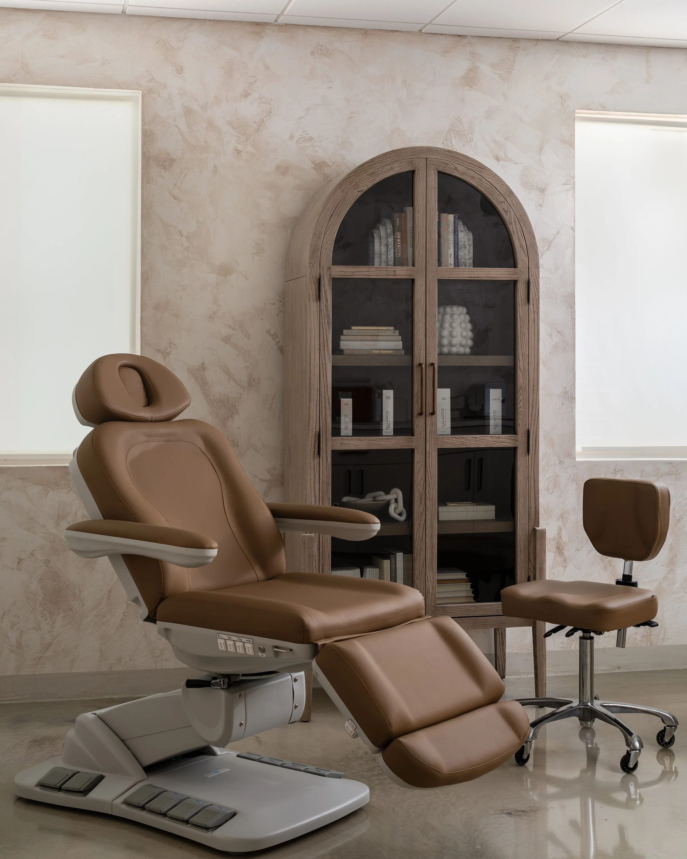 Medical examination room with an adjustable brown leather chair, a brown rolling stool, and a wooden glass-front cabinet against a beige textured wall.