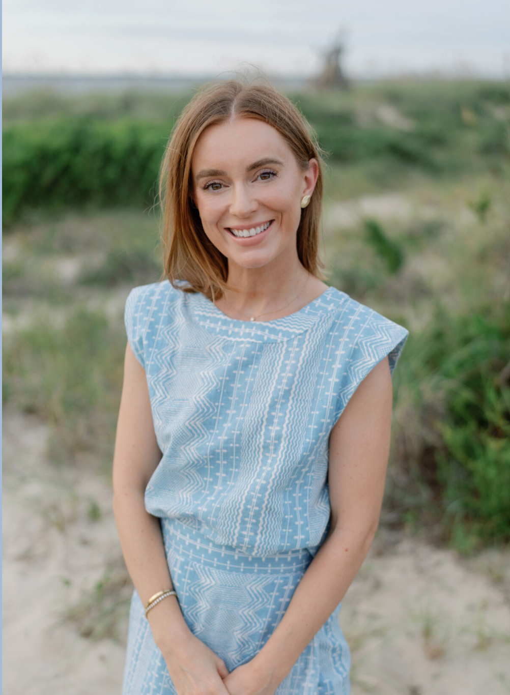A smiling woman with shoulder-length auburn hair wearing a sleeveless light blue dress with white geometric patterns, outdoors on a sandy and grassy terrain with greenery in the background.