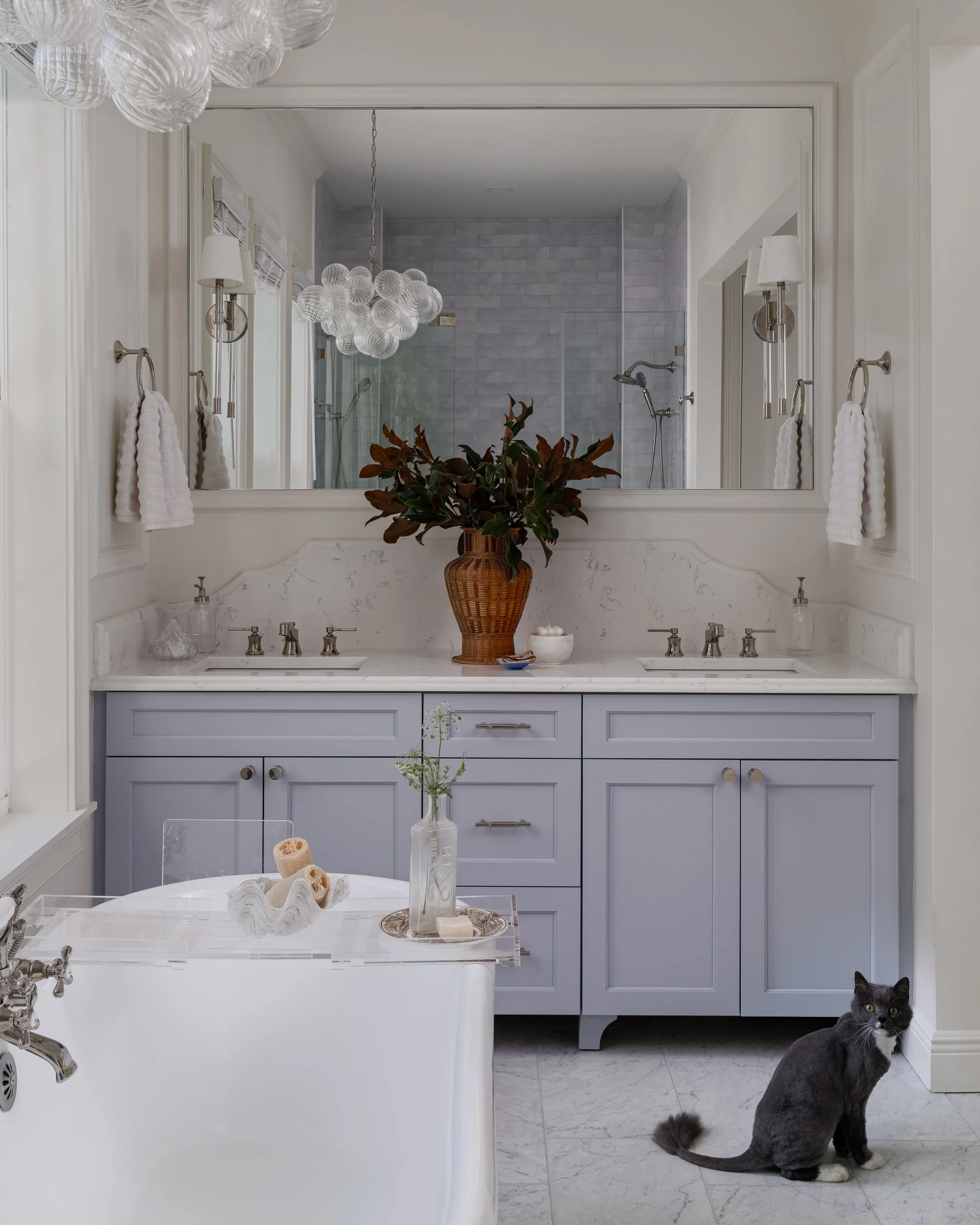 A bathroom with a double sink vanity, large mirror, glass chandelier, and a black cat sitting on the floor next to the bathtub.