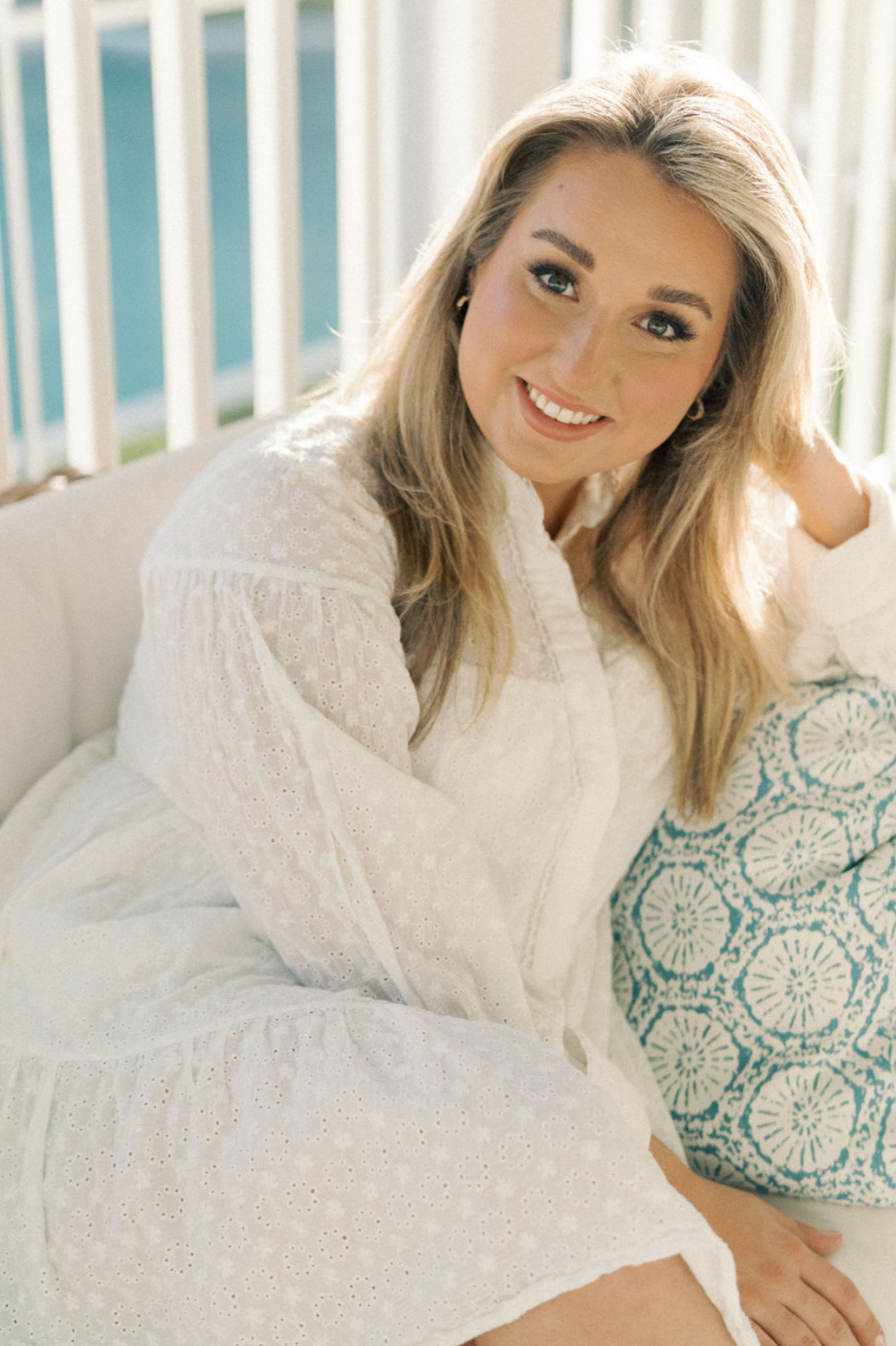 A woman with long blonde hair, smiling, sitting on a sofa with a patterned pillow in a well-lit room.