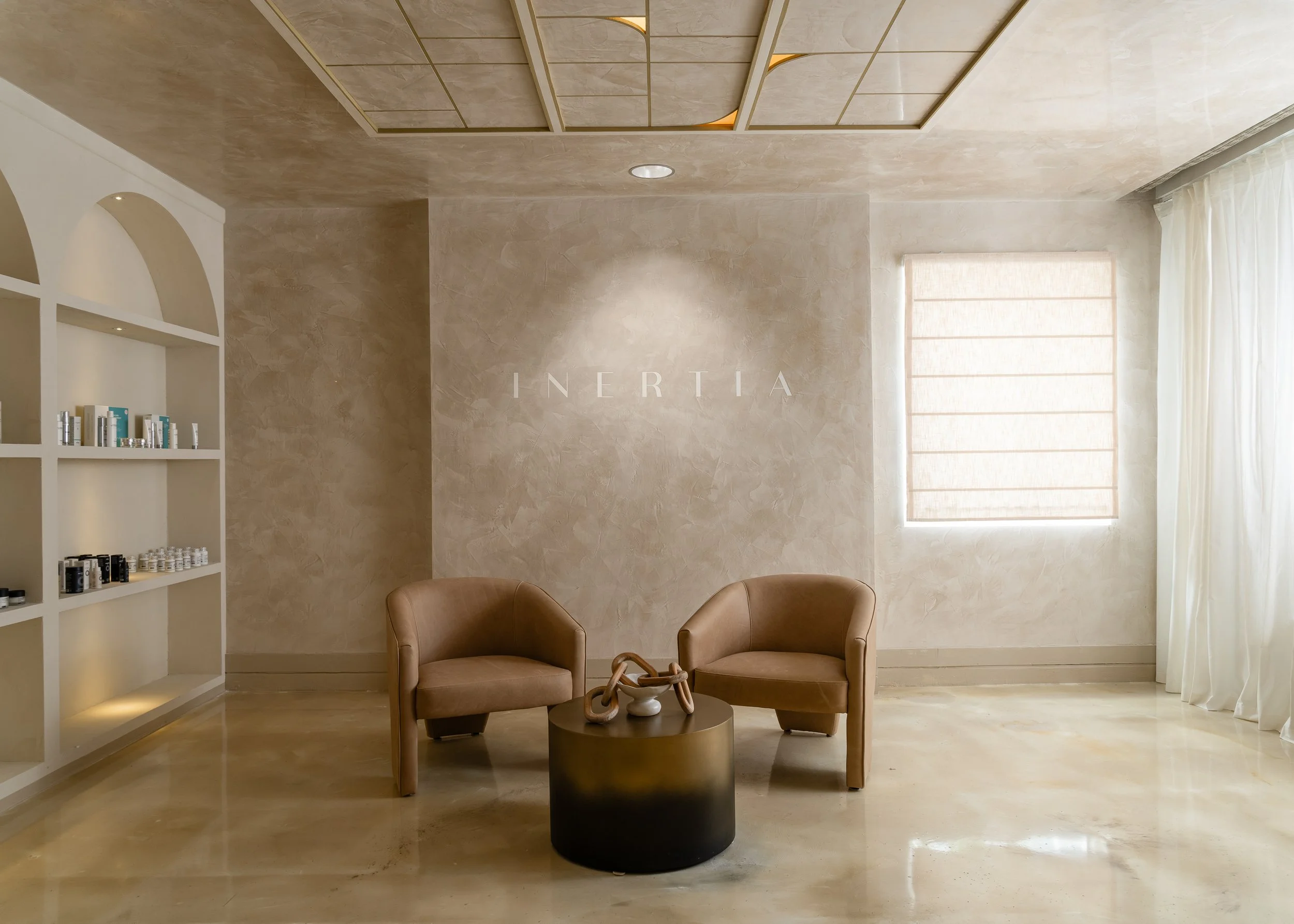 Minimalist beige lounge with two armchairs around a circular table decorated with a sculpture, a window with sheer curtains, and a wall with the white text 'INERTIA' on it.