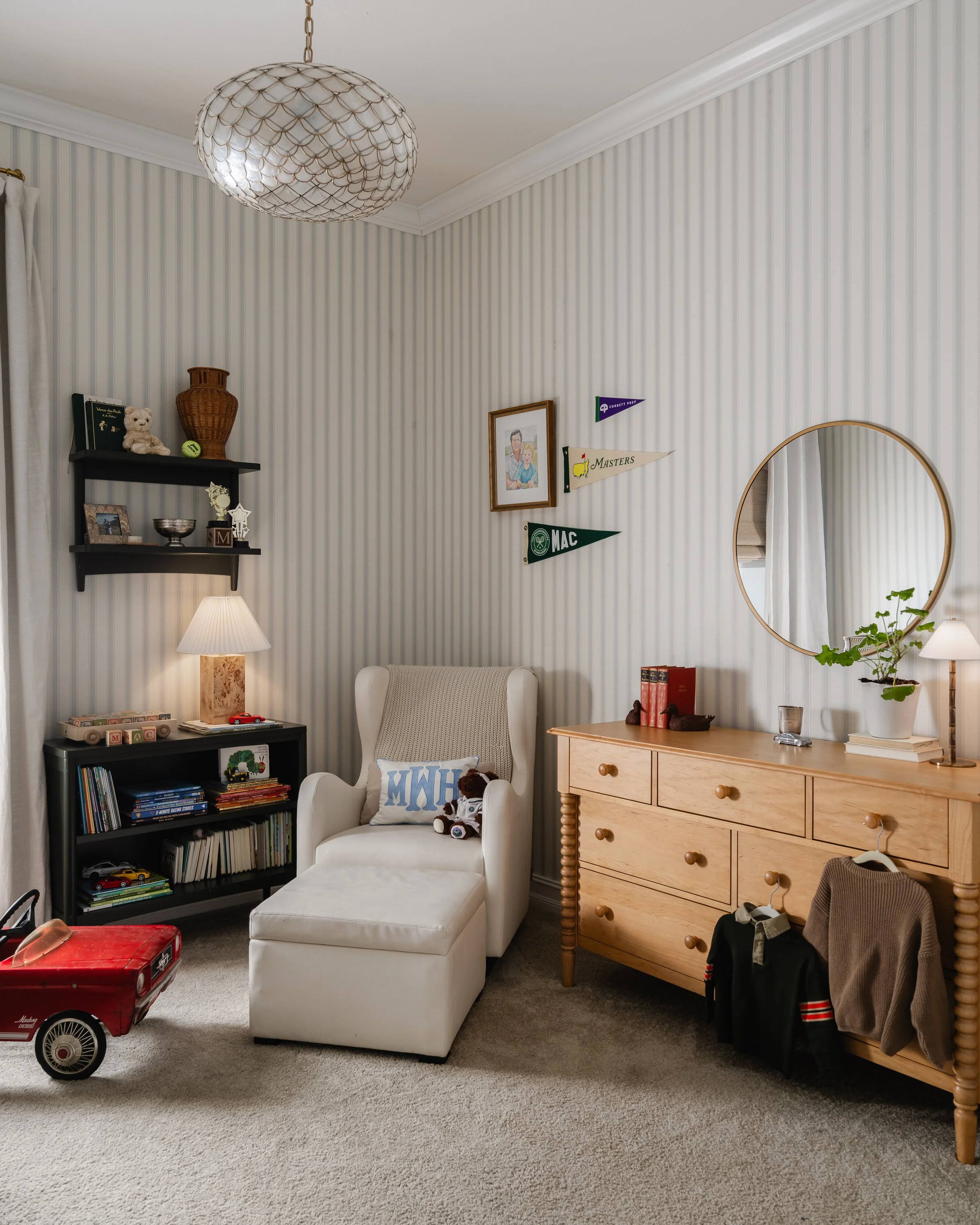 A cozy living room corner with a white armchair, a wooden dresser, a round mirror, a bookshelf, and decorative flags on the wall.