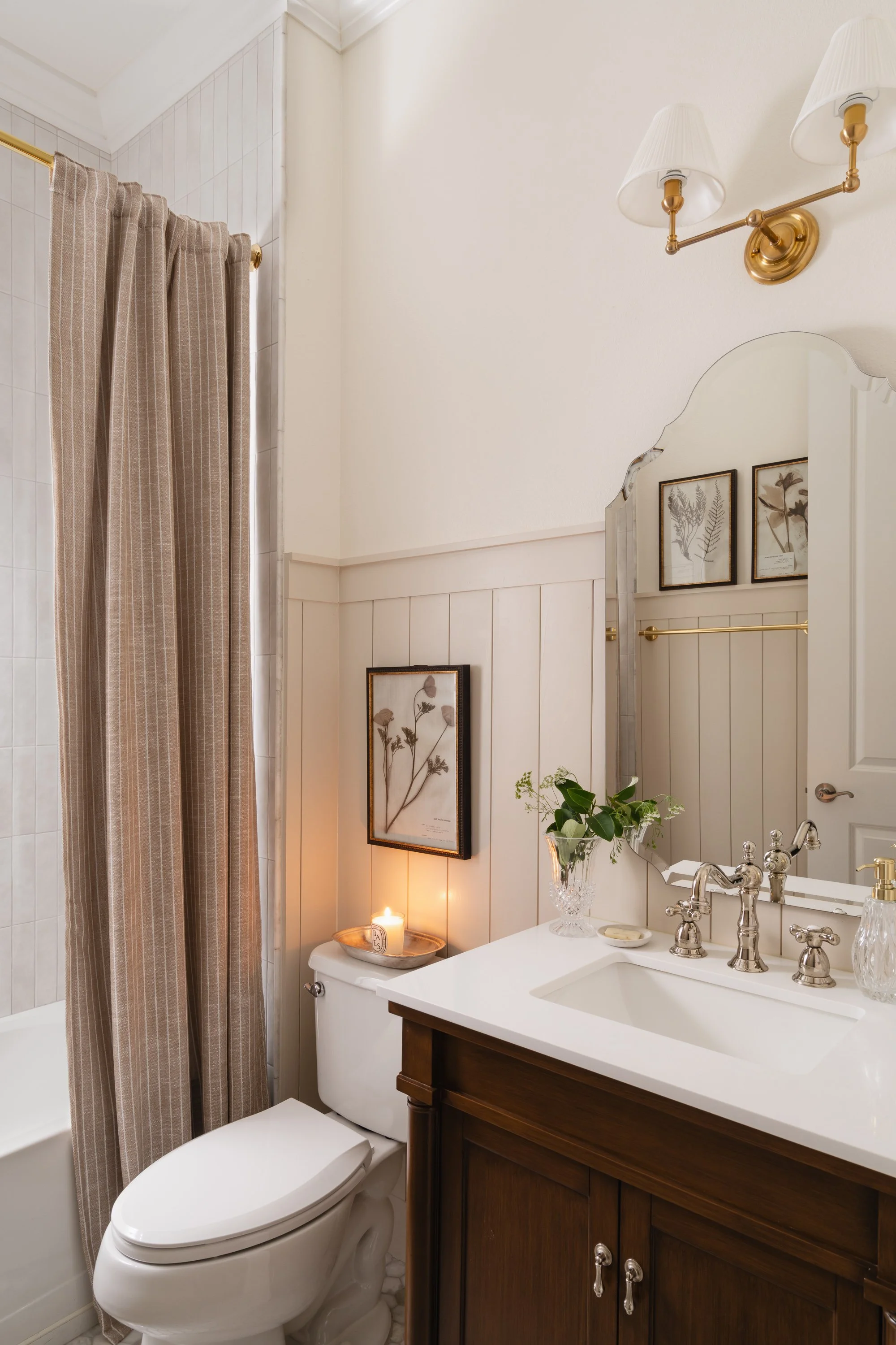 Bathroom with a white toilet, beige striped shower curtain, wooden vanity with white countertop, oval mirror, framed botanical prints, small lit candle, glass vase with greenery, and brass fixtures.