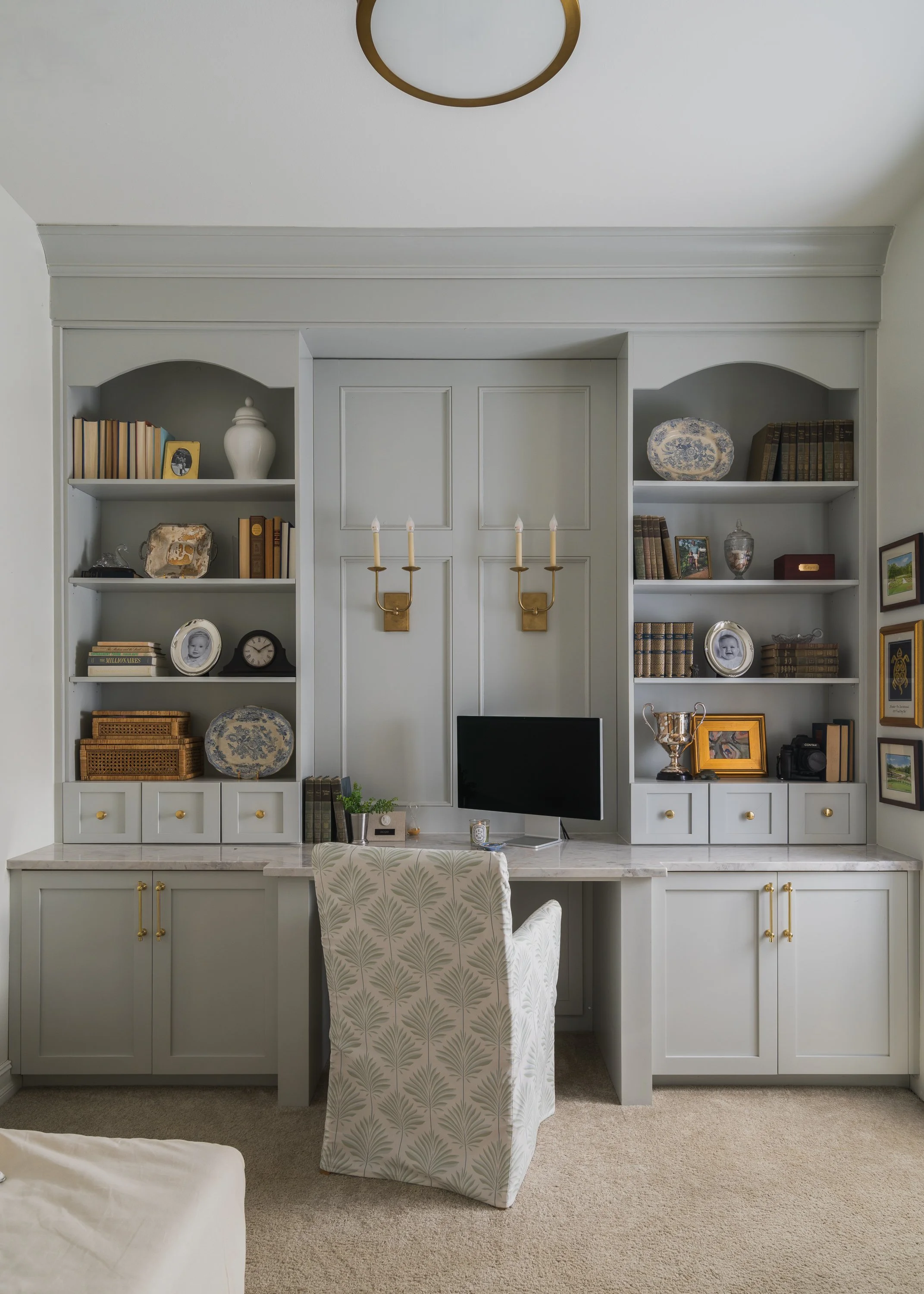 Home office with built-in white shelves filled with books, decorative items, and photo frames. A desk with a chair, a flat-screen monitor, and wall-mounted gold candle sconces.