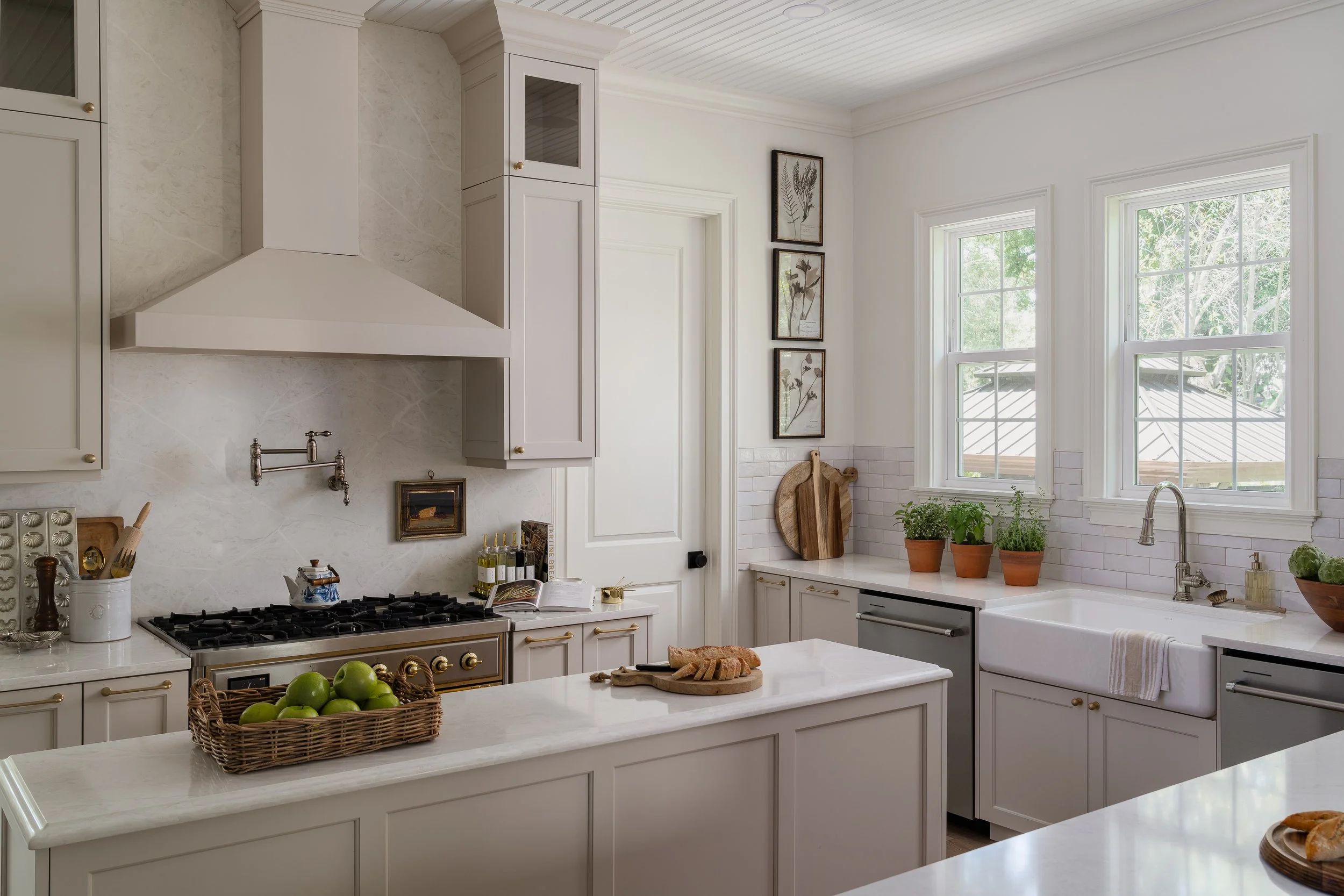 Bright kitchen with white cabinets, marble countertops, and a farmhouse sink. There are two large windows with greenery outside, potted plants, wooden cutting boards, and a basket of green apples on the island.