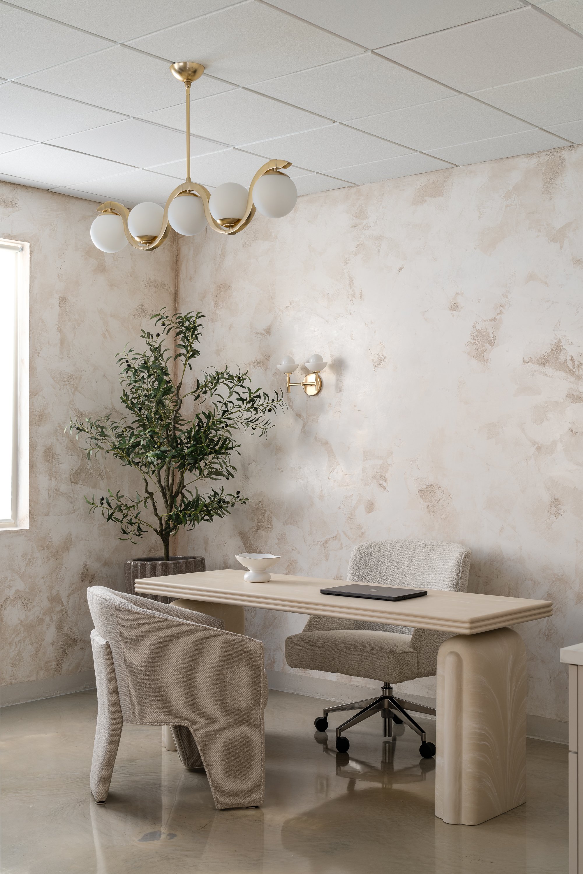 A minimalistic office or meeting room with a cream-colored desk, two upholstered chairs, a potted plant, modern wall and ceiling light fixtures, and a window allowing natural light.