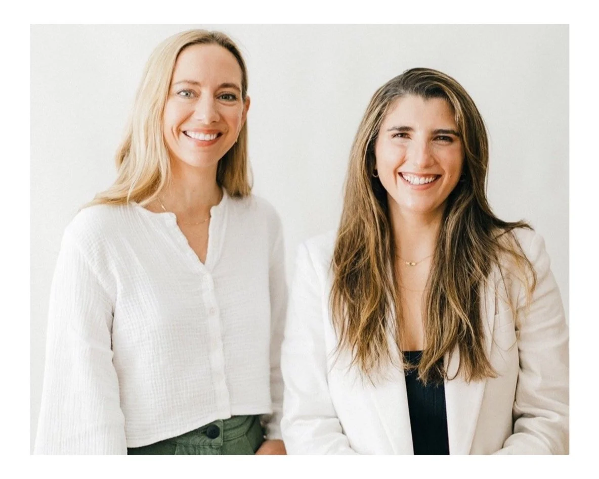 Two women standing side by side, smiling against a plain white wall. The woman on the left has blonde hair and wears a white blouse, while the woman on the right has long brown hair, wears a white blazer over a black top, and has gold jewelry.