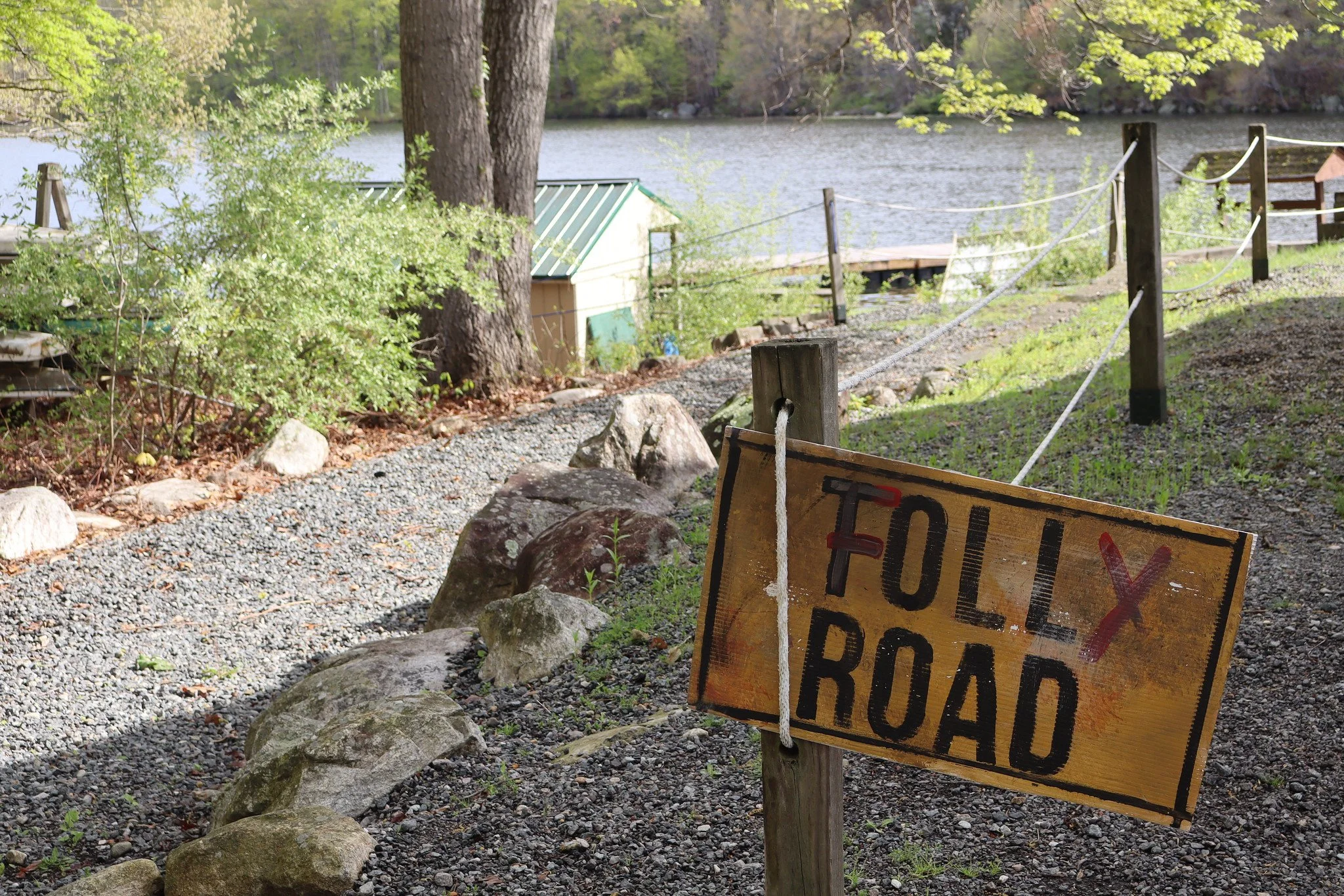 A lakeside scene with trees, a dock, a small shed, and a rusty warning sign in the foreground that is partially obscured.