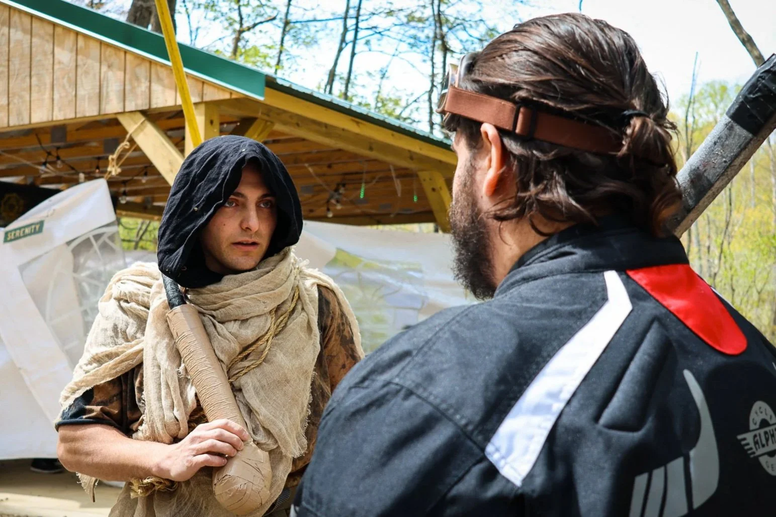 Two men having a serious conversation outdoors, one wearing a black hood and beige scarf, holding a makeshift weapon, and the other with glasses and long hair, wearing a black jacket with red and white accents, behind a wooden structure surrounded by trees.