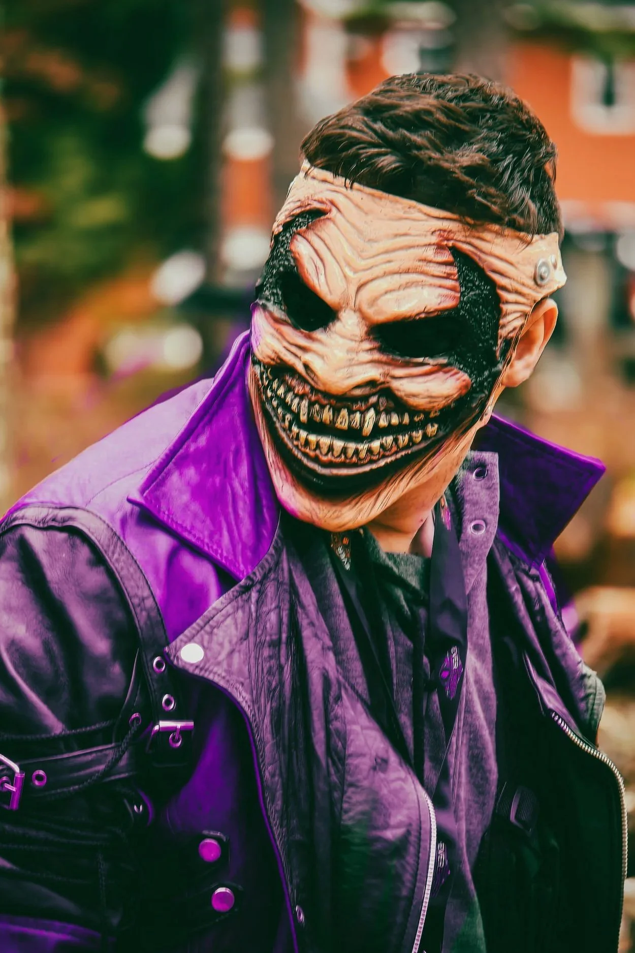 Person wearing a zombie mask with twisted, grotesque features, black eyes, and a wide, jagged smile, dressed in a purple leather jacket and black clothing, outdoors with blurred background