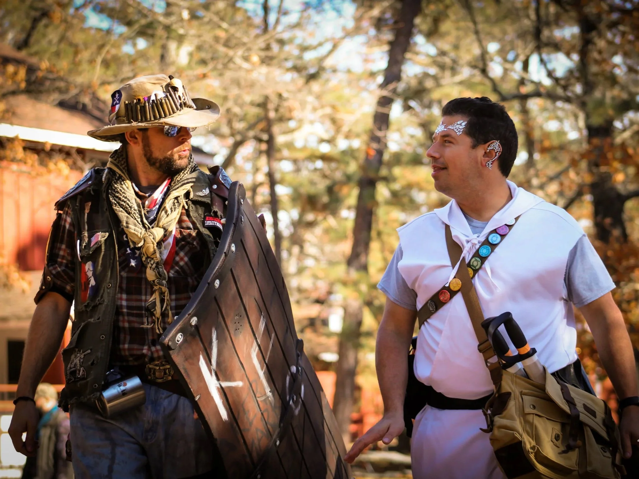 Two men, one dressed in military or tactical gear with a large hat, sunglasses, and a flag bandana, and the other in casual clothing with accessories, are engaged in conversation outdoors on a sunny fall day with trees in the background.