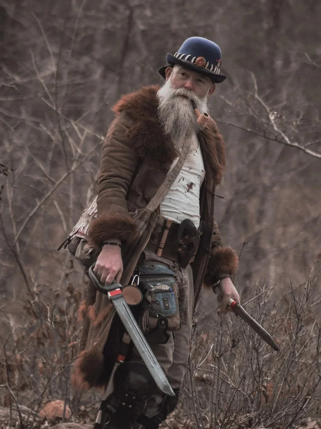 A man dressed as a hunter with a long gray beard, wearing a blue hat decorated with feathers and a brown fur coat, standing in a wooded area holding a knife and a machete.