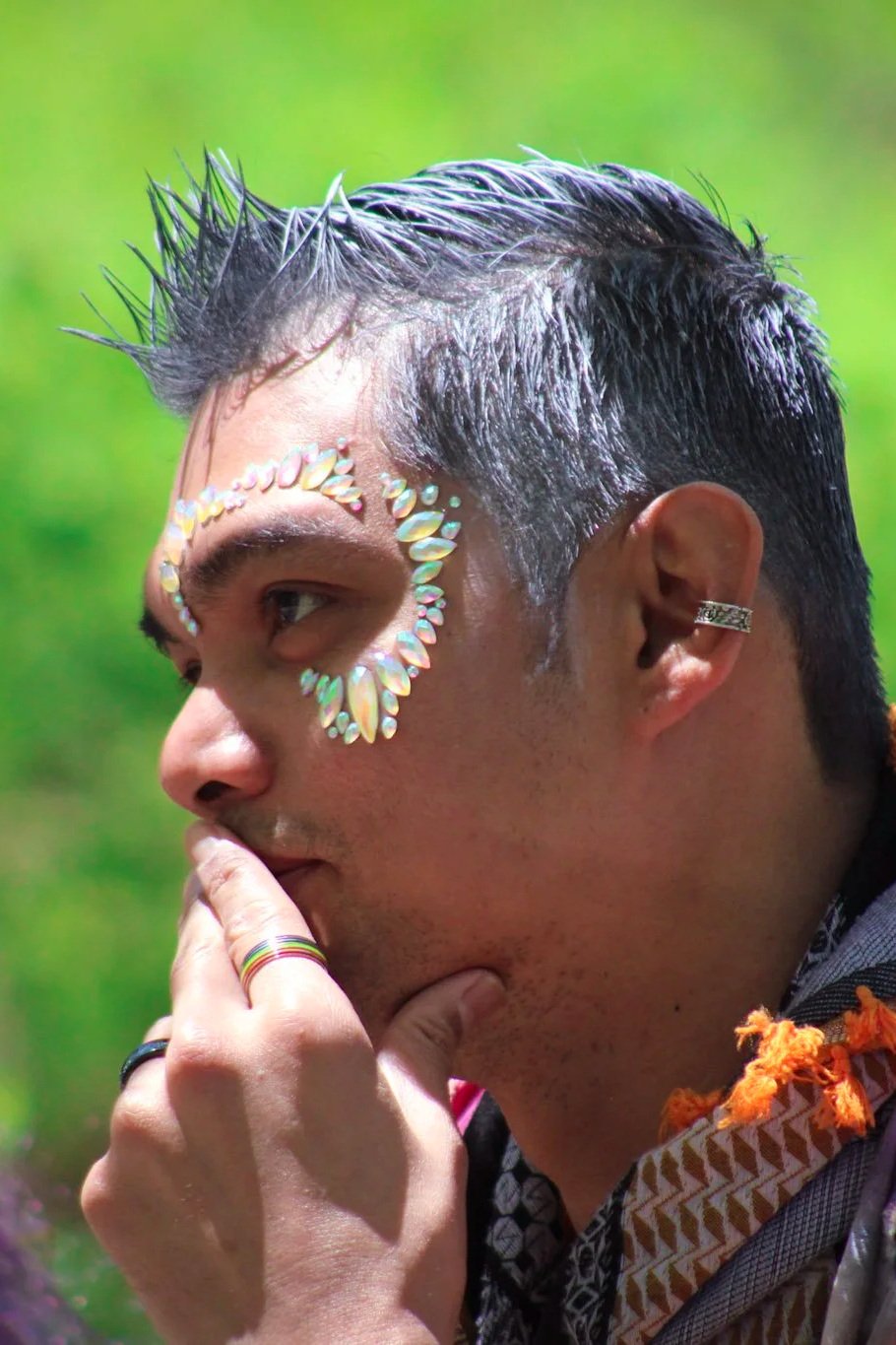 A person with short, slicked-back black hair and decorative gemstones on their face, standing outdoors with a green blurred background. They are touching their lips with their hand, wearing rings and an earring, and dressed in patterned clothing with orange accents.
