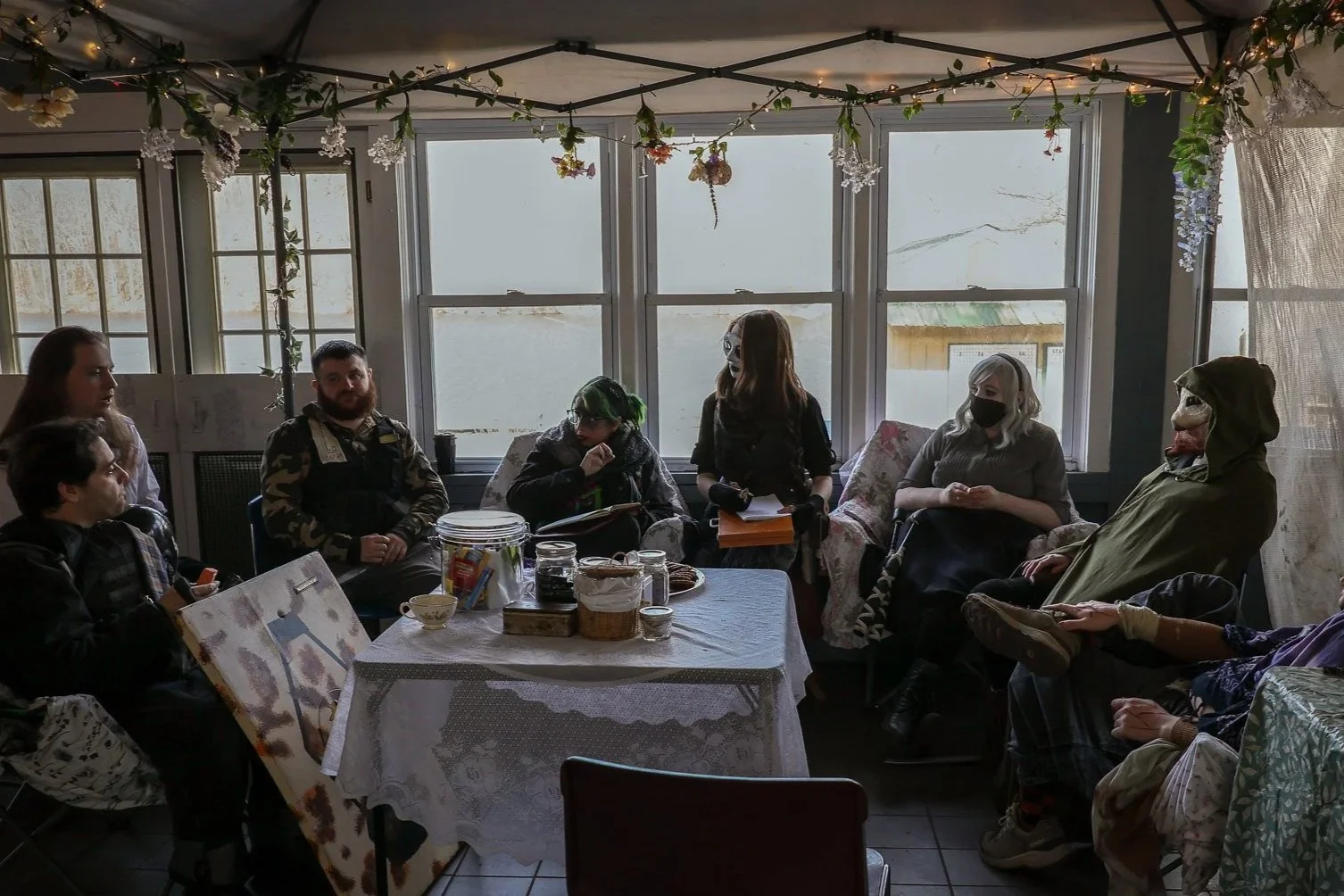 Group of people sitting around a table in a room decorated with hanging flowers, some wearing masks and costumes, engaged in a discussion or meeting.