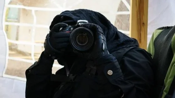 Person in black outdoor gear taking a photo with a camera, seated in a wooden and fabric structure.