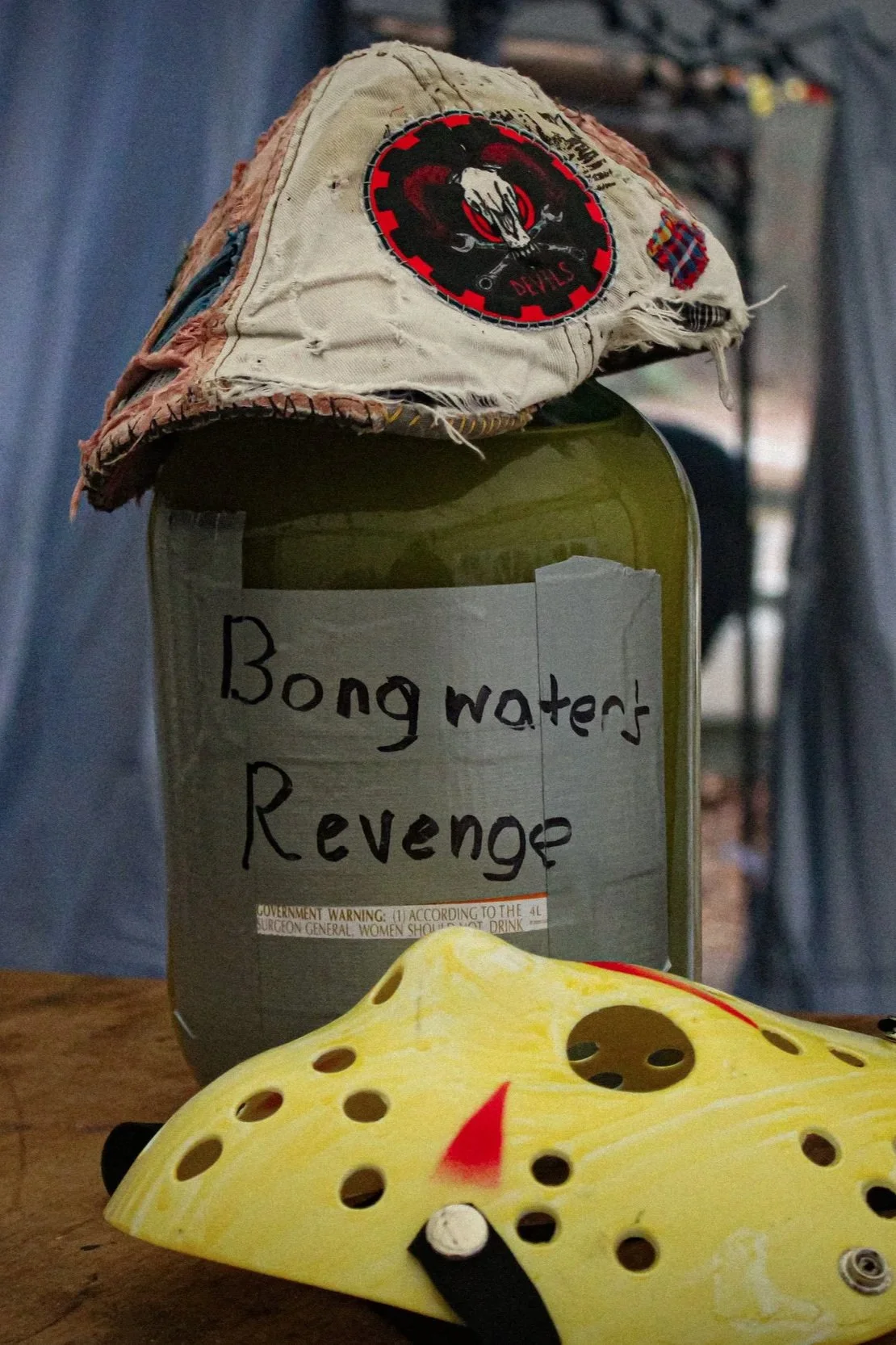 A plastic mask of a yellow mouse with holes and painted facial features, placed in front of a canister labeled 'Bong water's Revenge' with a worn cloth hat on top, which has a patch with a skull and crossed scalpels design.