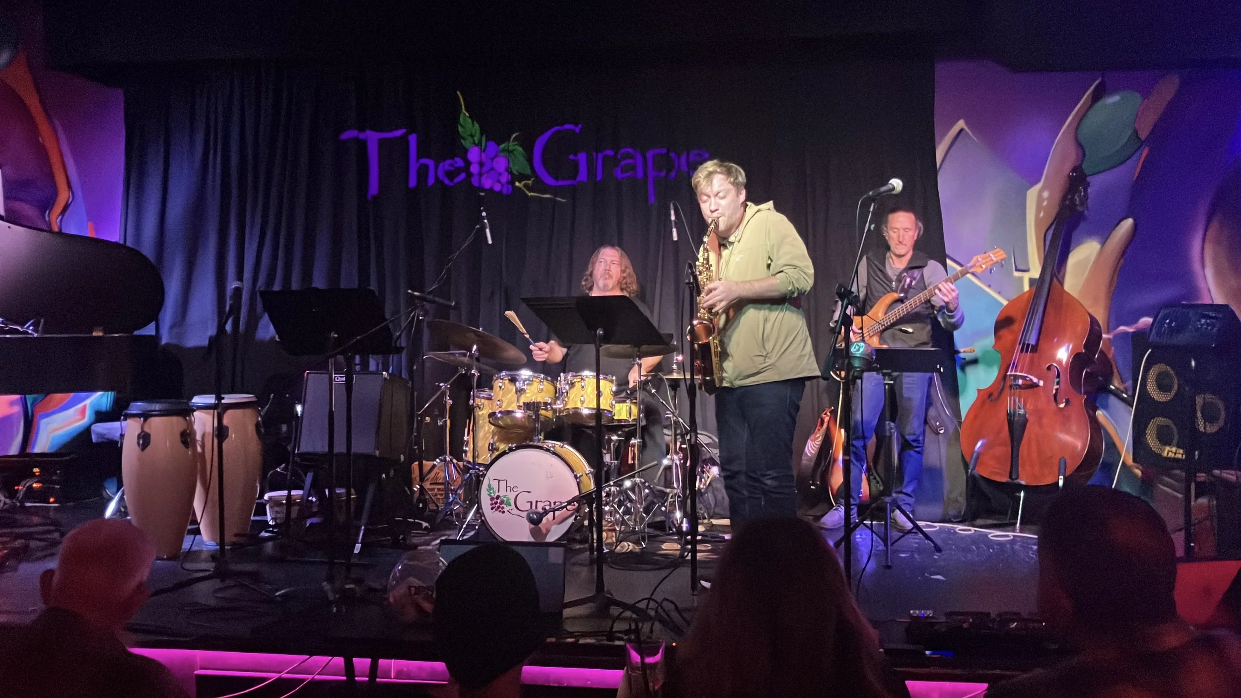 A jazz band performs on stage at The Grape. The band includes a drummer, a saxophonist, a bassist, and a pianist. The stage is decorated with a colorful mural in the background and the venue's sign, "The Grape," is visible above the performers.