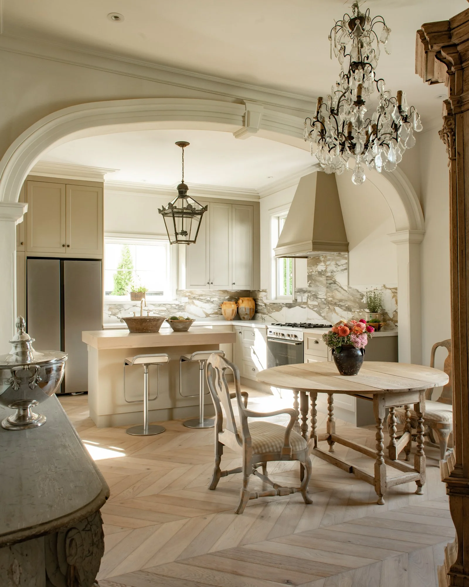 Elegant kitchen with cream-colored cabinets, marble backsplash, wooden dining table with floral centerpiece, vintage chairs, chandelier, and pendant light.